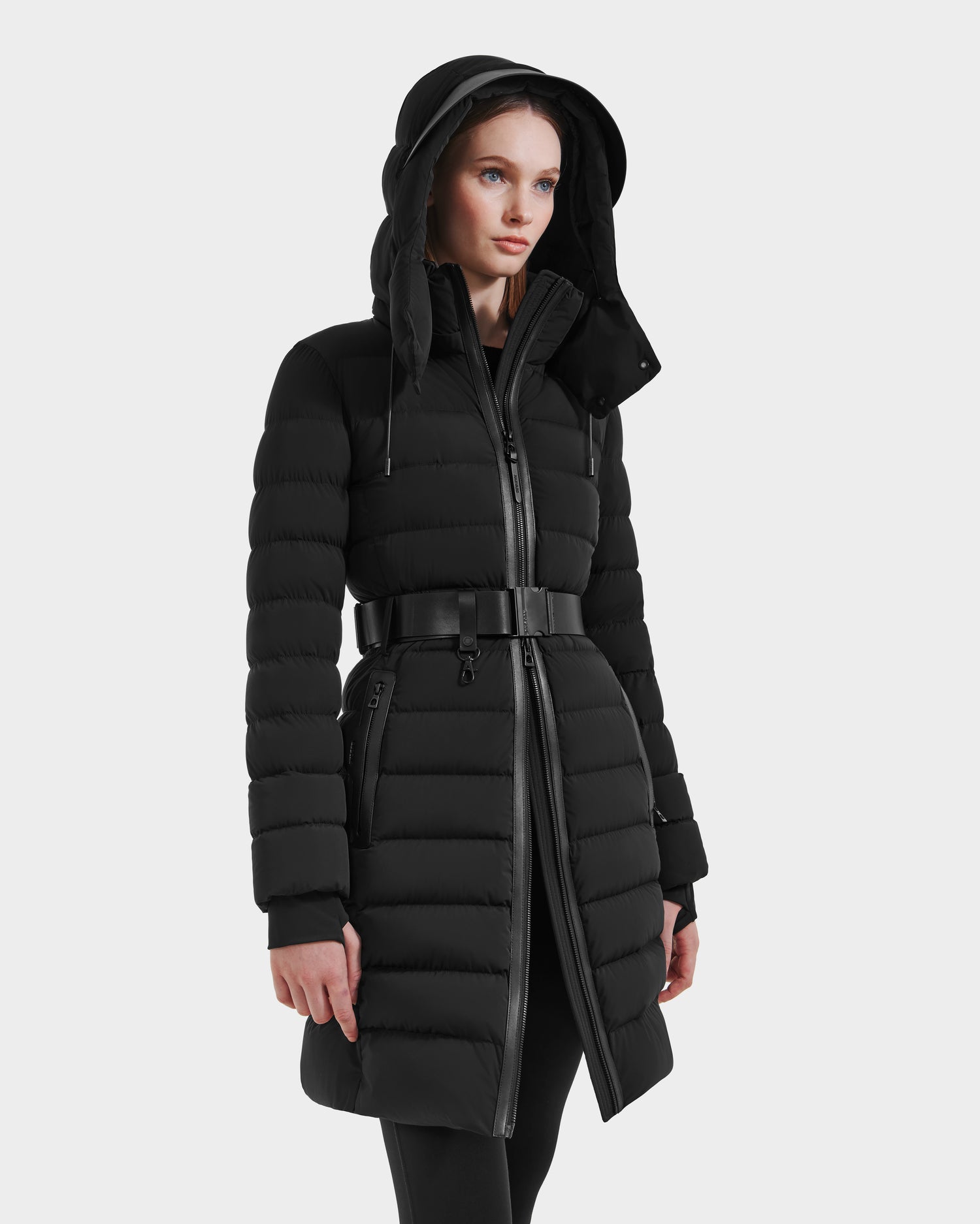 A woman wears the Rudsak PELIN M HV - H125508 Black: a long, black belted down jacket with a hood, front zipper, and horizontal quilted pattern, standing against a plain white background.