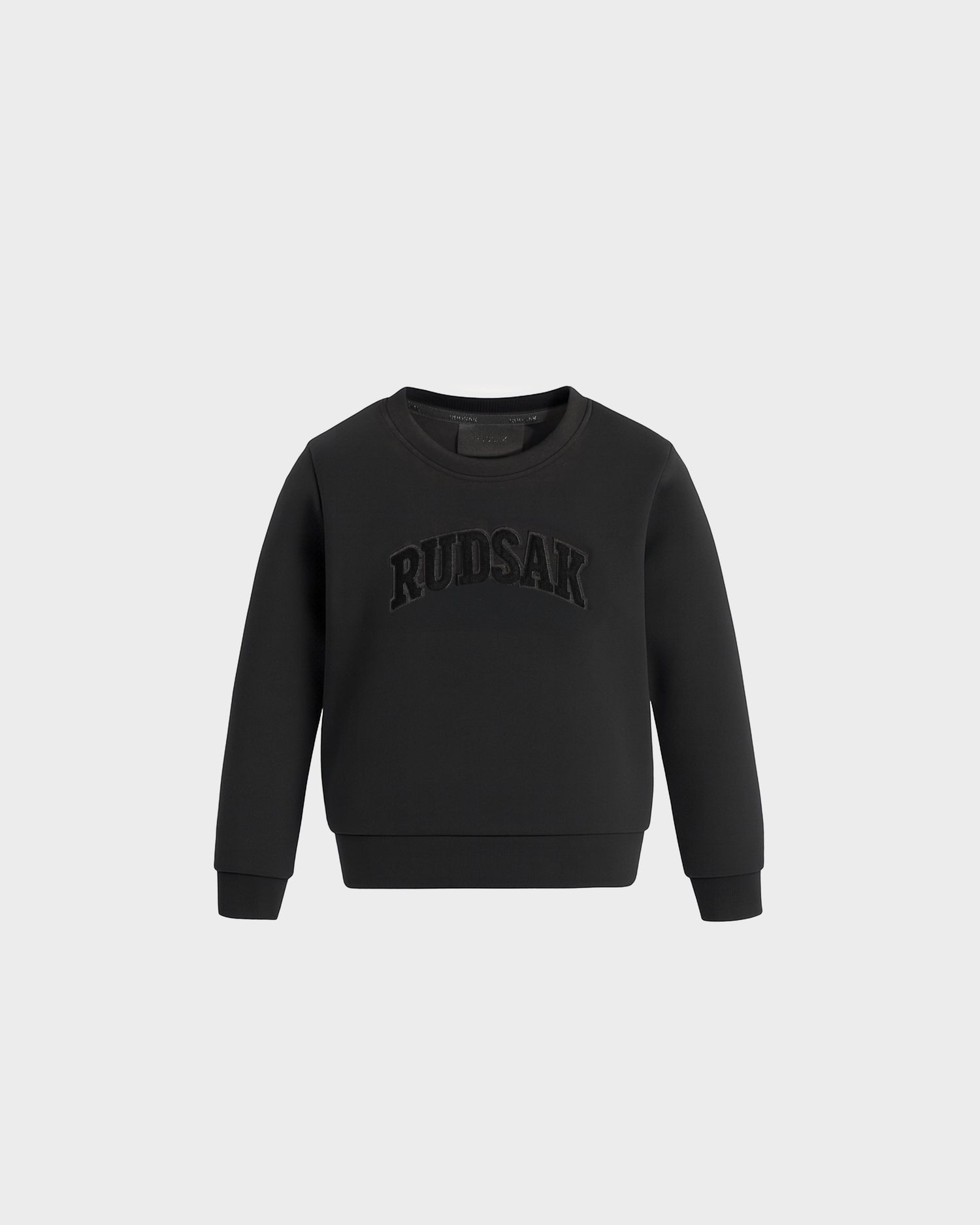 The Rudsak POPPY - 1525003 Black is a black long-sleeve sweatshirt featuring bold “RUDSAK” embroidery across the chest, shown against a plain light gray background.