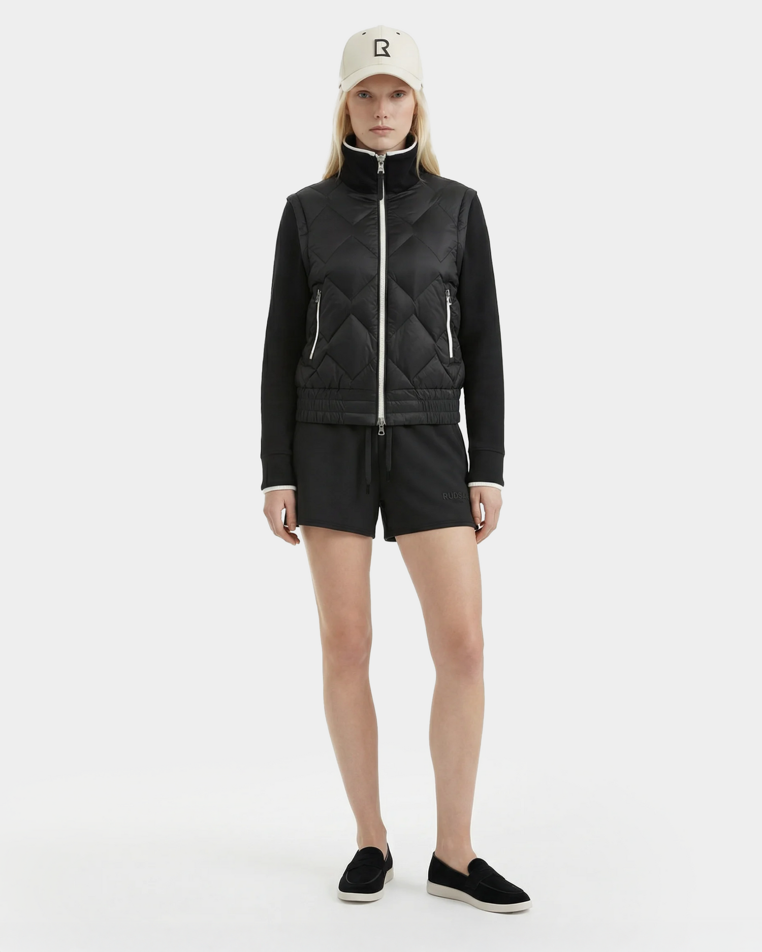 A woman faces forward against a white background, wearing the Rudsak PRISA - 8126022 Black convertible down jacket with removable sleeves, paired with black shorts, black slip-on shoes, and a beige baseball cap featuring a black R.
