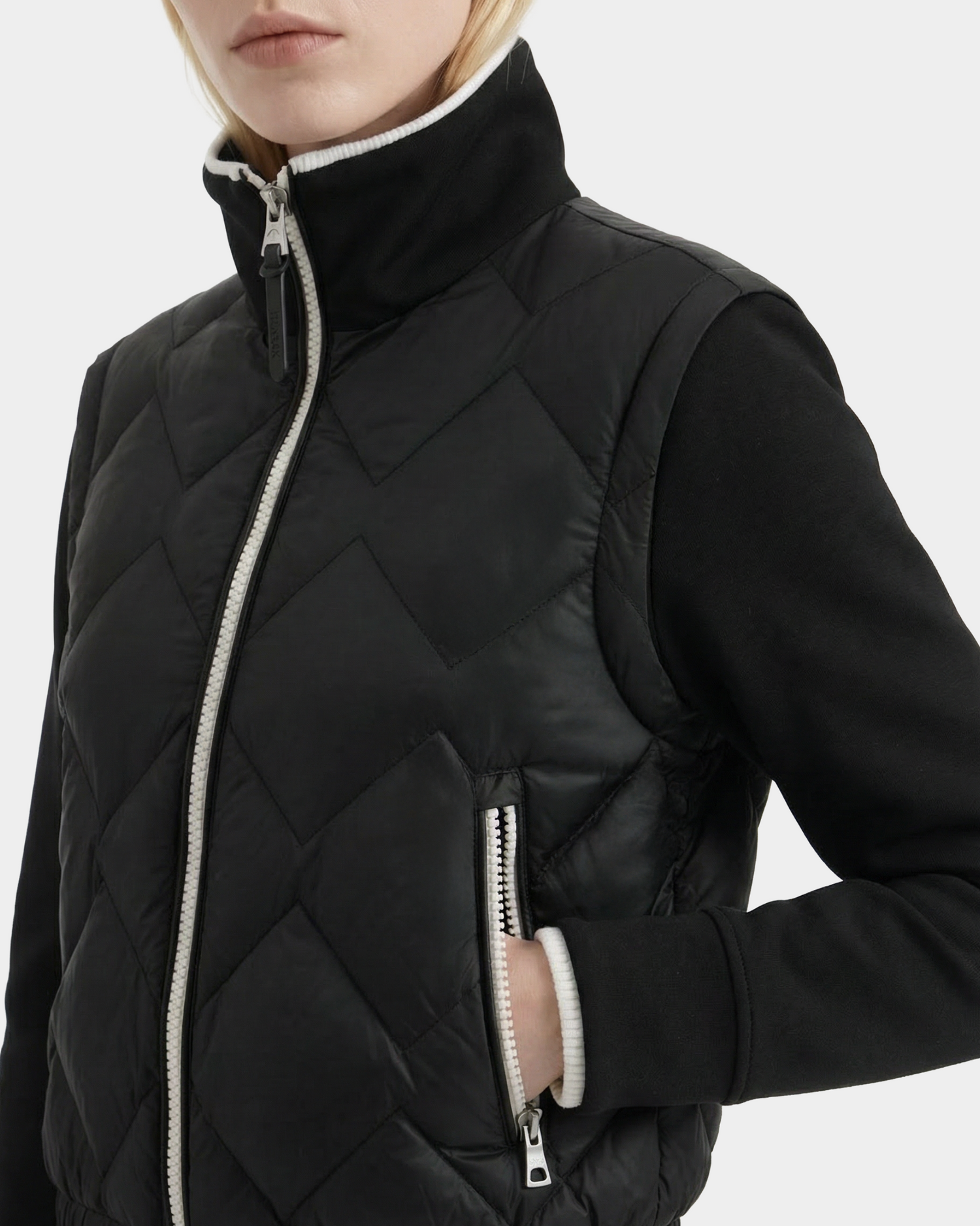 A person wearing the Rudsak PRISA - 8126022 Black quilted vest with a high collar and white zipper over a black long-sleeve top, styled like a convertible down jacket with removable sleeves, shown from shoulders to waist.