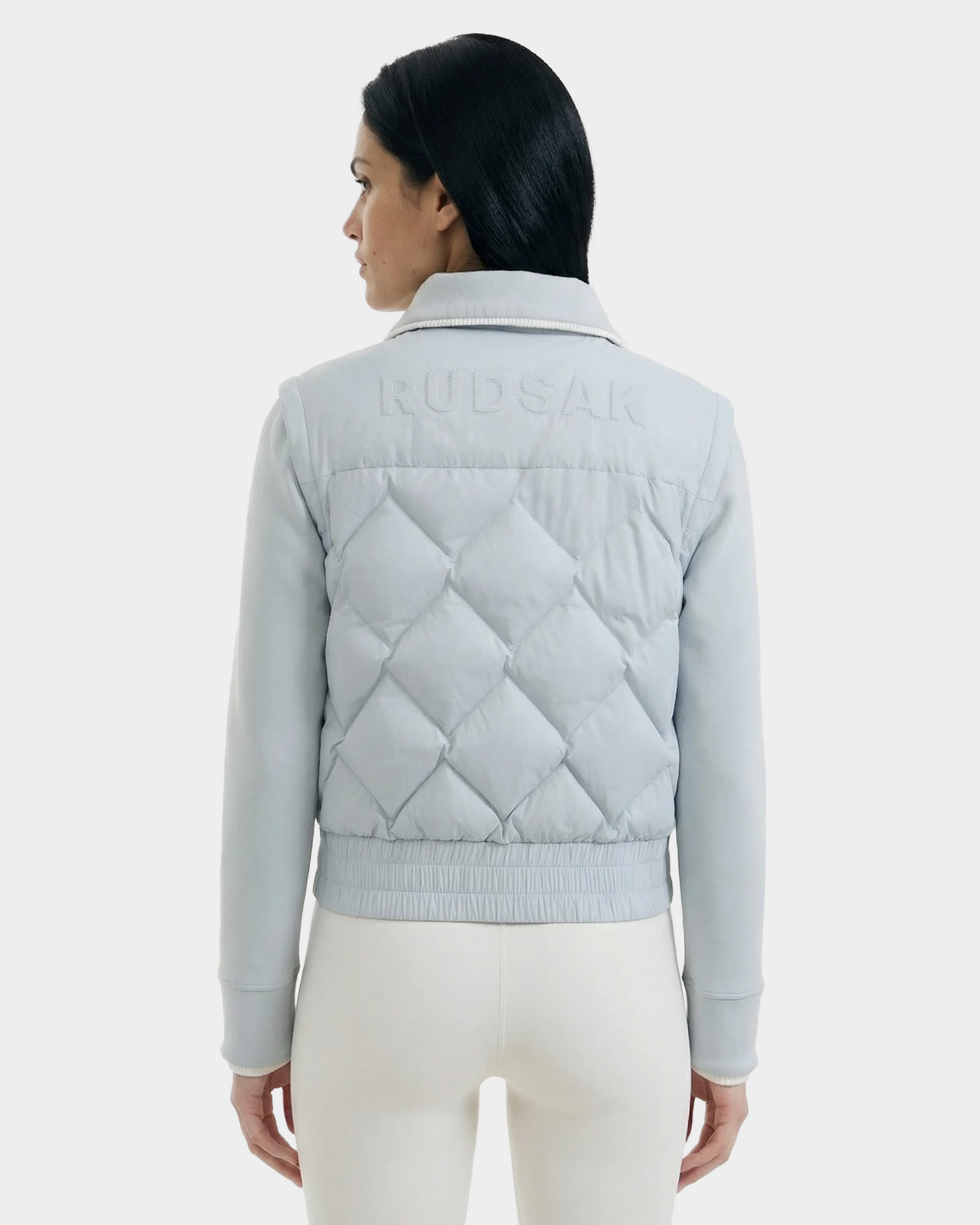 A woman with long dark hair, seen from behind, wears the Rudsak PRISA - 8126022 Powder Blue convertible down jacket and light-colored pants, standing against a plain white background.