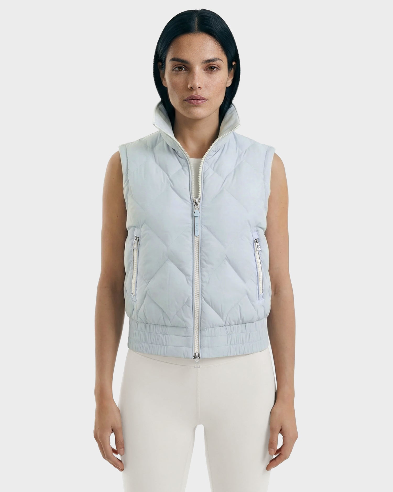 A woman with straight dark hair wears the Rudsak PRISA - 8126022 Powder Blue convertible jacket with removable sleeves and a high collar over a light outfit, standing against a plain, light background.