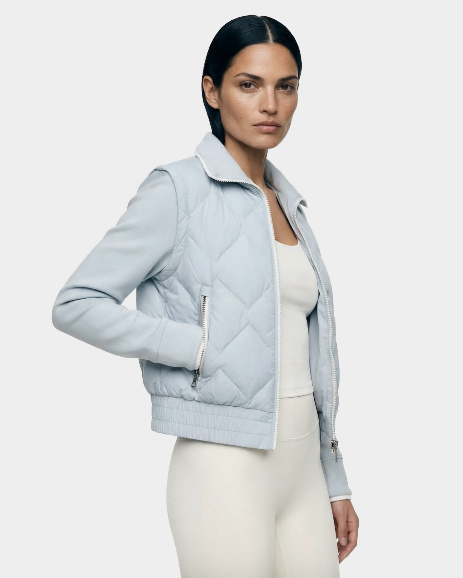 A woman with straight dark hair wears the Rudsak PRISA - 8126022 Powder Blue convertible down jacket over a white top and white pants, standing against a plain light gray background.