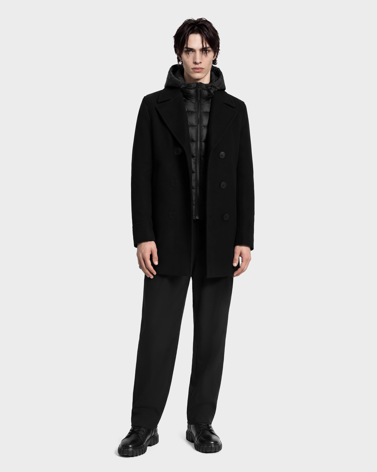 A person wears a black LUCIEN - 6126508 Black double-breasted pea coat over a black quilted hooded puffer vest, black trousers, and black lug-sole shoes.