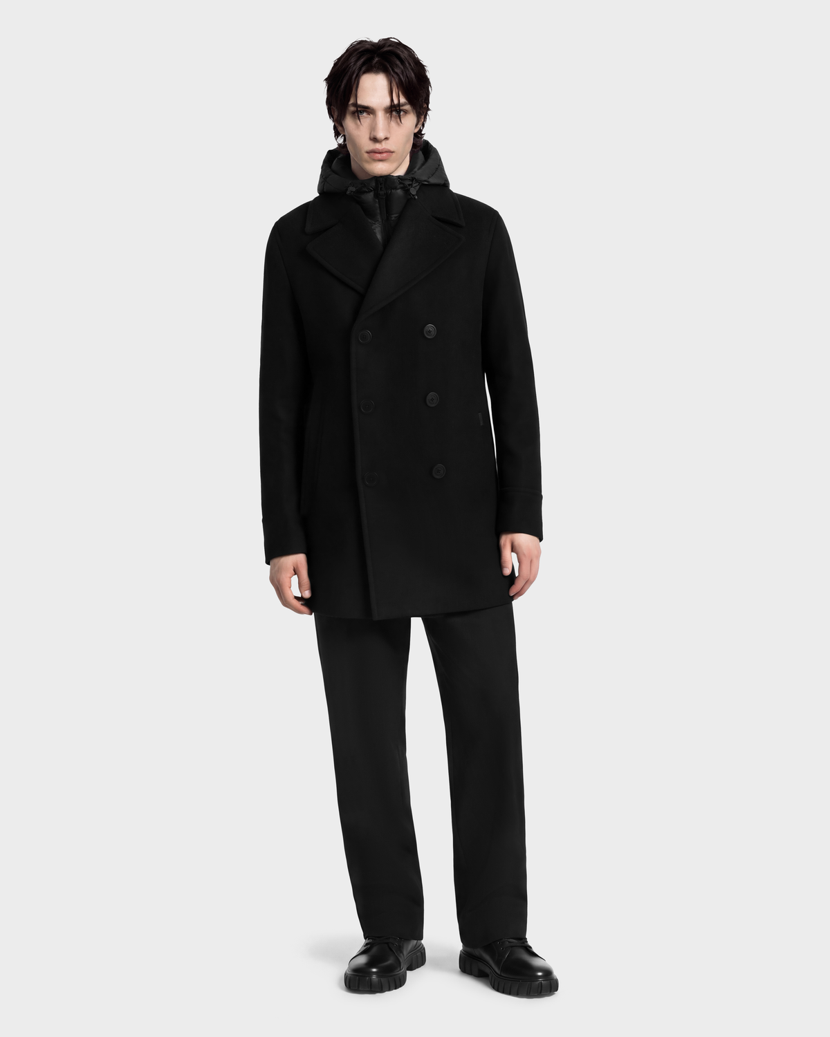 A person wears a black LUCIEN - 6126508 Black double-breasted pea coat layered over a black hooded jacket and black trousers.