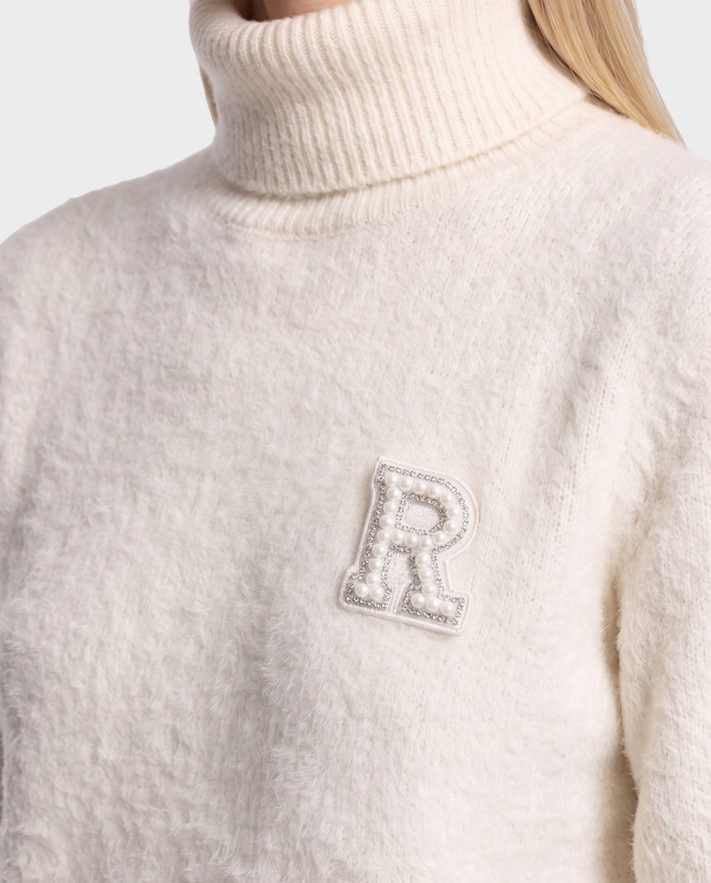 Wearing the ALISYA - 8526505 Iceland by Rudsak, a cream-colored fuzzy knit turtleneck sweater with a relaxed silhouette and a pearl-embellished letter R patch on the chest. The person’s face is not visible.
