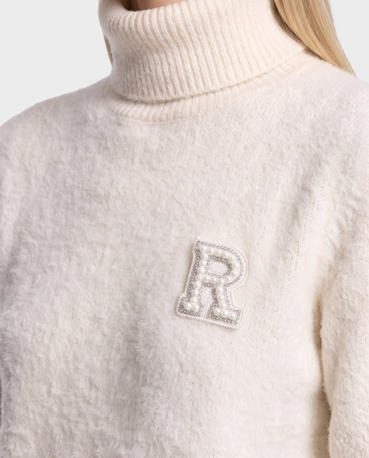 Wearing the ALISYA - 8526505 Iceland by Rudsak, a cream-colored fuzzy knit turtleneck sweater with a relaxed silhouette and a pearl-embellished letter R patch on the chest. The person’s face is not visible.