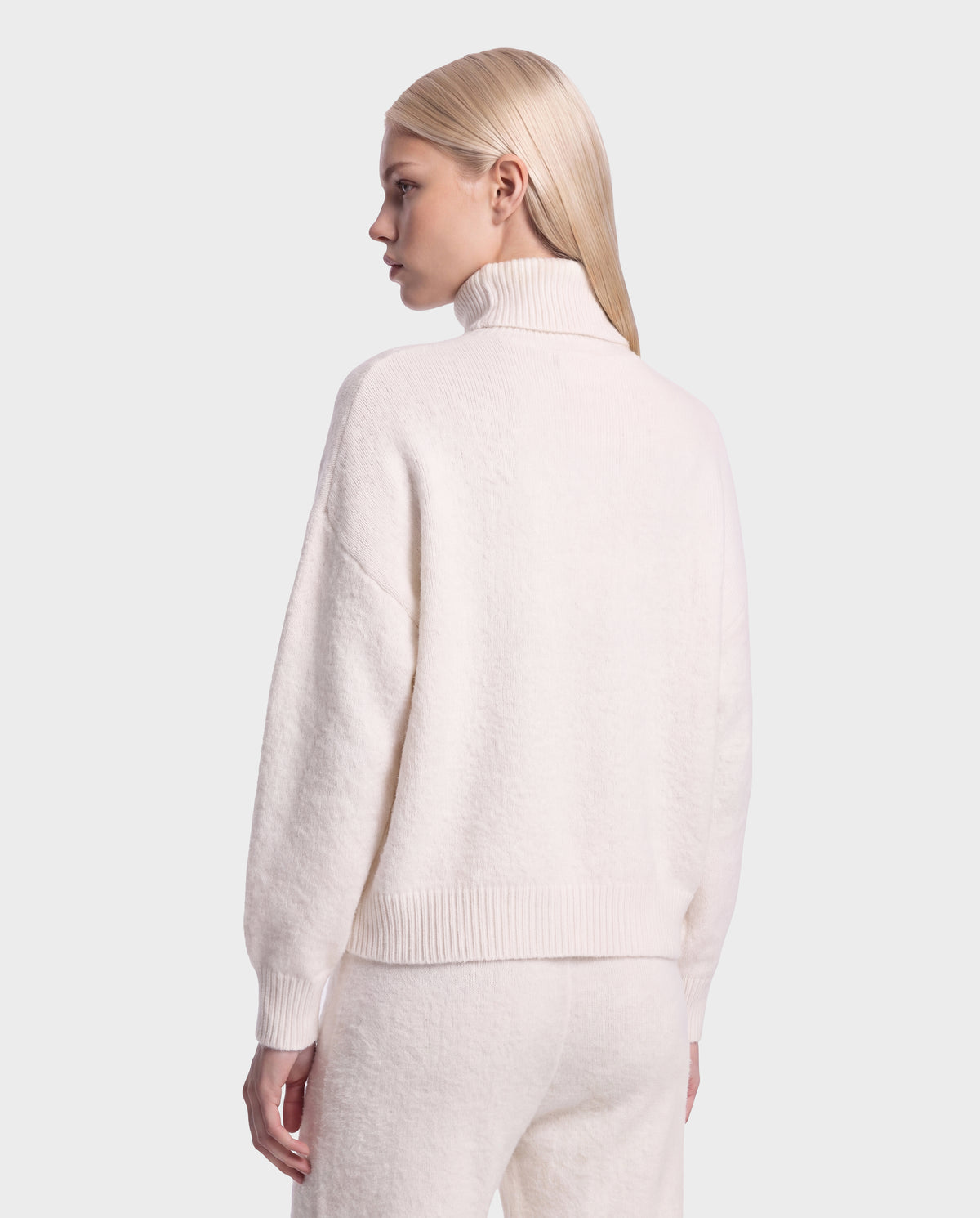 A person with straight, blonde hair is seen from behind, wearing the Rudsak ALISYA - 8526505 Iceland fuzzy knit turtleneck sweater and matching pants in a relaxed silhouette, set against a plain light background.