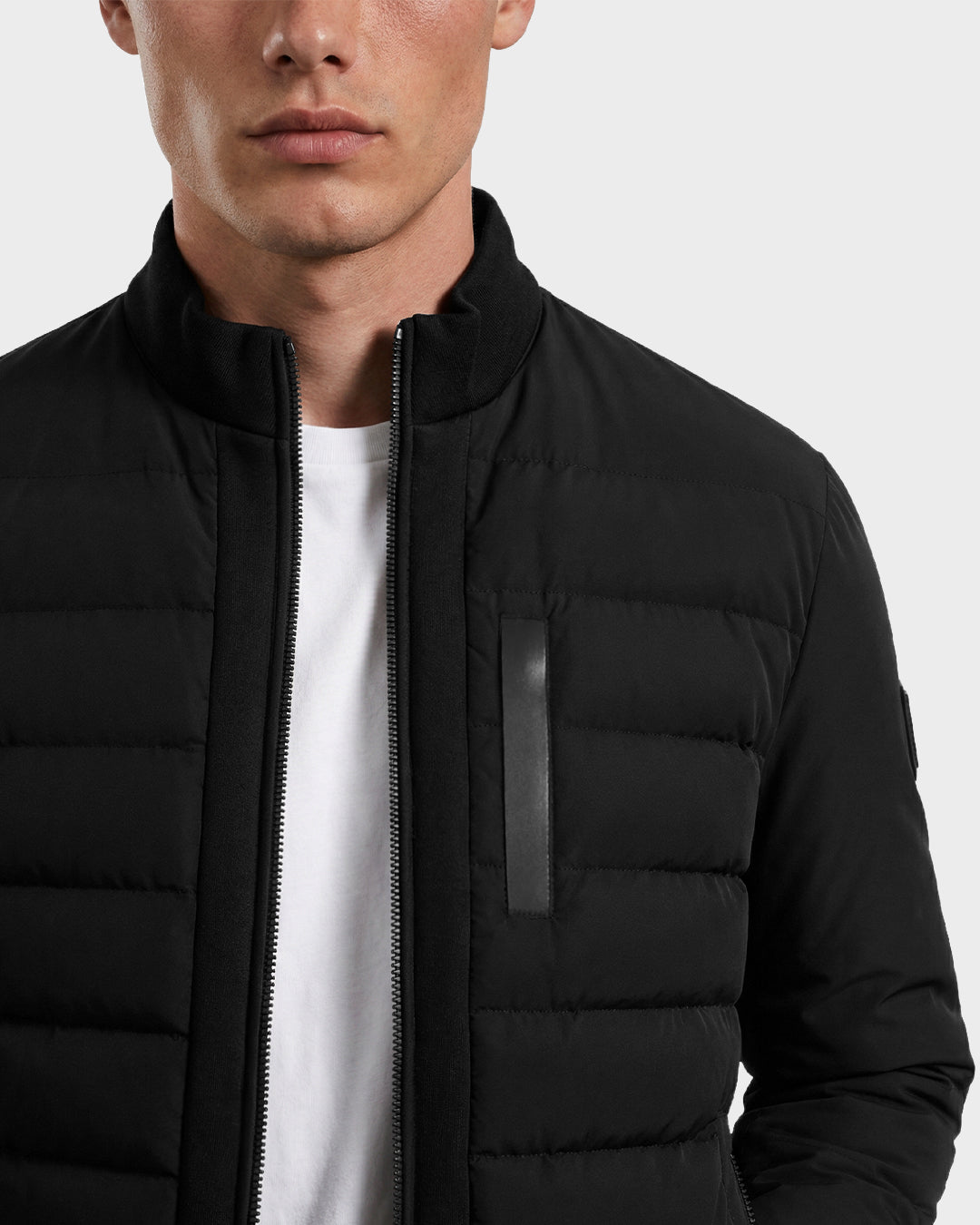 A man wearing the Rudsak JIMMY - 6125723 Black men's quilted down jacket, water and wind-resistant, over a white T-shirt, is shown from shoulders to just above the mouth against a plain light background.