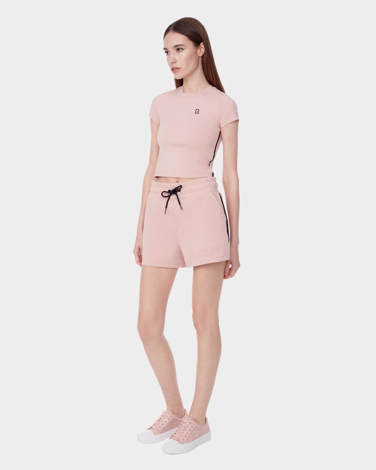 A woman with long brown hair stands against a white background wearing Rudsak’s ISLA - 8523000 BLUSH neoprene shorts, a light pink fitted crop top, and pink sneakers, reflecting Rudsak's modern style.