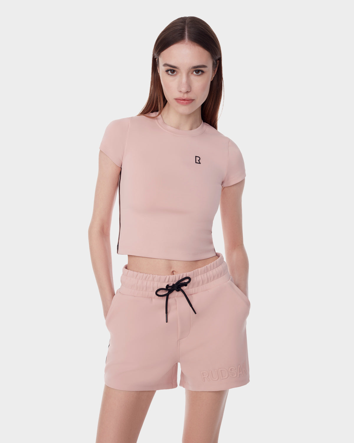 A woman stands with hands in pockets against a white background, wearing the Rudsak ISLA - 8523000 BLUSH fitted crop top and matching drawstring shorts, both crafted from sleek neoprene scuba fabric.