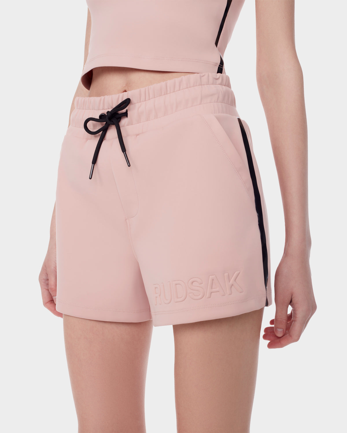 A person wears Rudsak's ISLA - 8523000 BLUSH light pink neoprene shorts, featuring a black drawstring, side stripe, and embossed logo on the left leg, paired with a matching blush crop top that's partially visible.