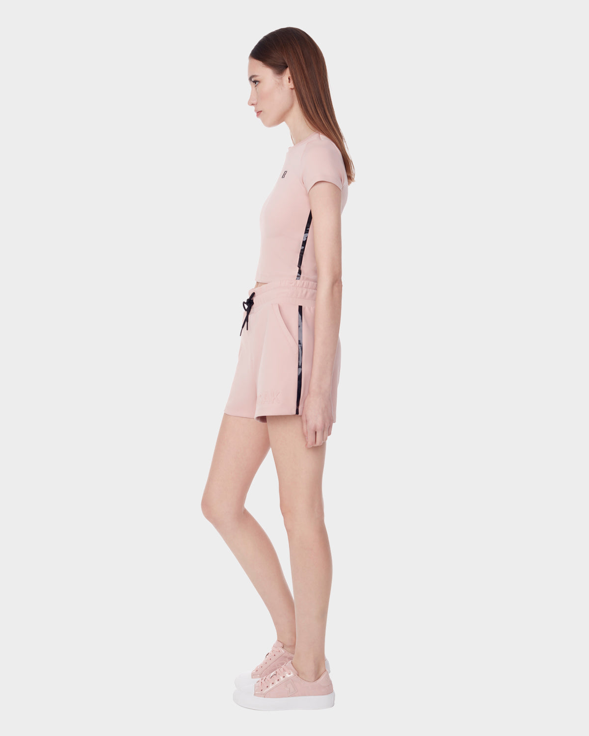 A woman stands in profile against a white background, wearing the Rudsak ISLA - 8523000 BLUSH shorts and a light pink short-sleeve top, both in neoprene scuba fabric, styled with pink sneakers and black drawstring accents.