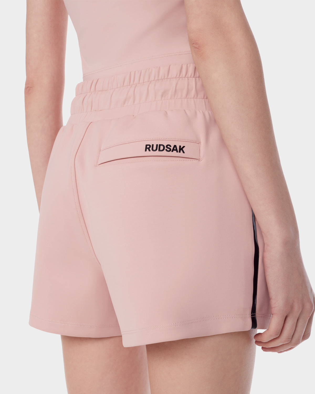 A person shown from the waist down, photographed from behind, wears Rudsak ISLA - 8523000 BLUSH shorts in light pink neoprene scuba fabric with an elastic waistband and a back pocket featuring the Rudsak label in black.