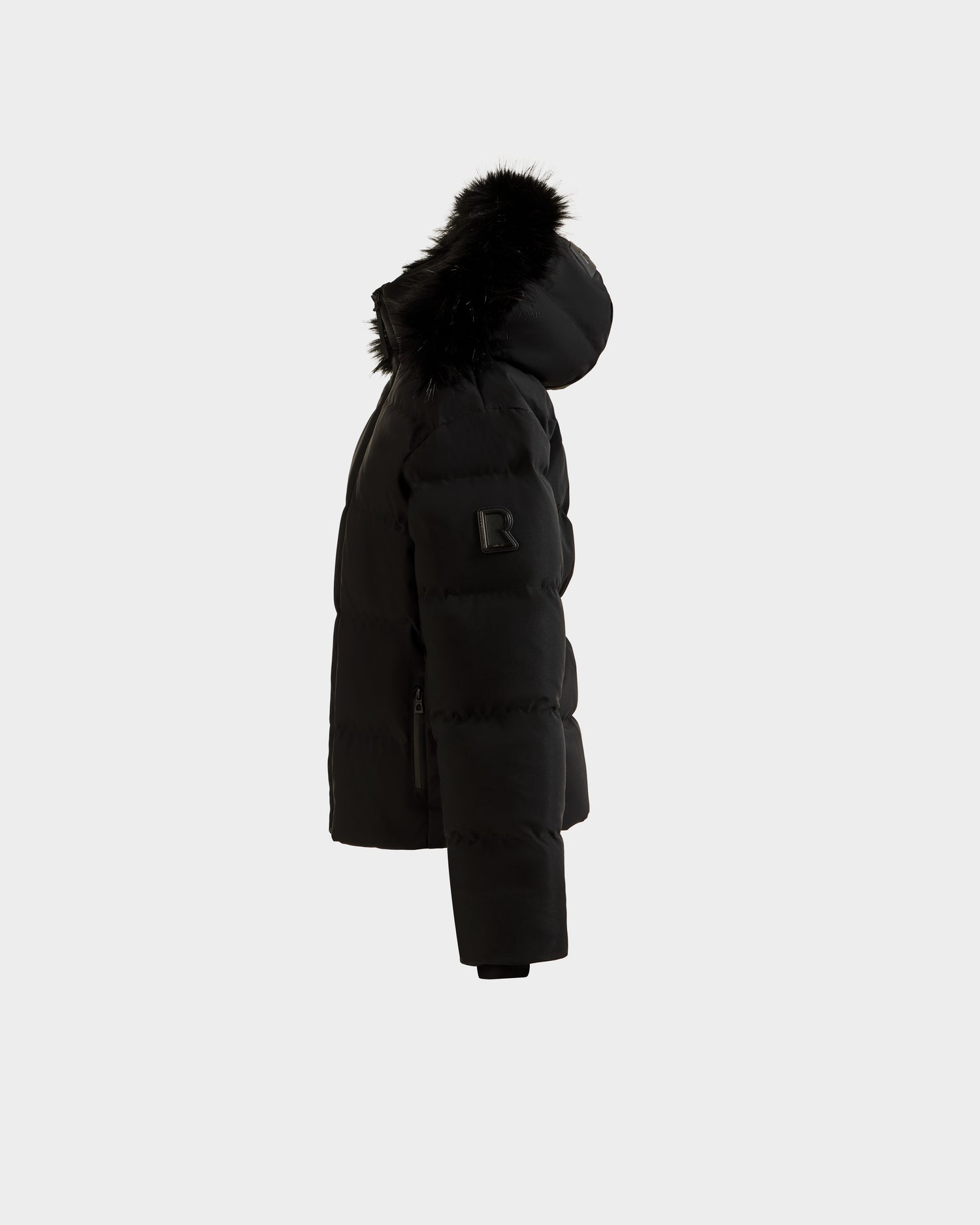 The Rudsak RAEGAN - 0125503 Black puffer jacket features a fur-trimmed hood and a sleeve patch, shown in side profile against a plain white background. The jacket appears warm and padded.
