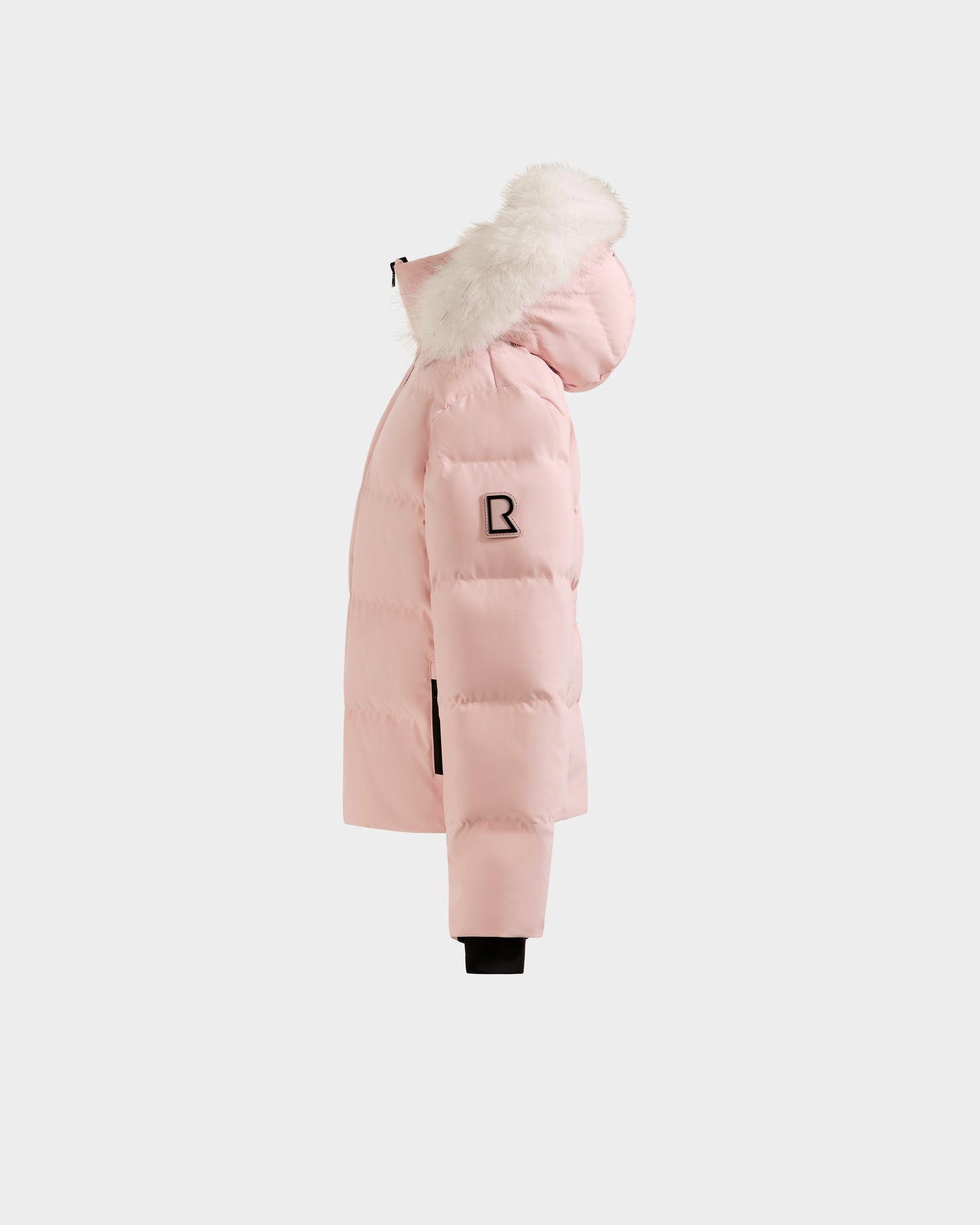 The Rudsak RAEGAN - 0125503 Pastel Pink is a light pink puffer jacket with a hood trimmed in white faux fur, black zipper details, and a black patch logo on the sleeve, shown in side profile against a plain white background.