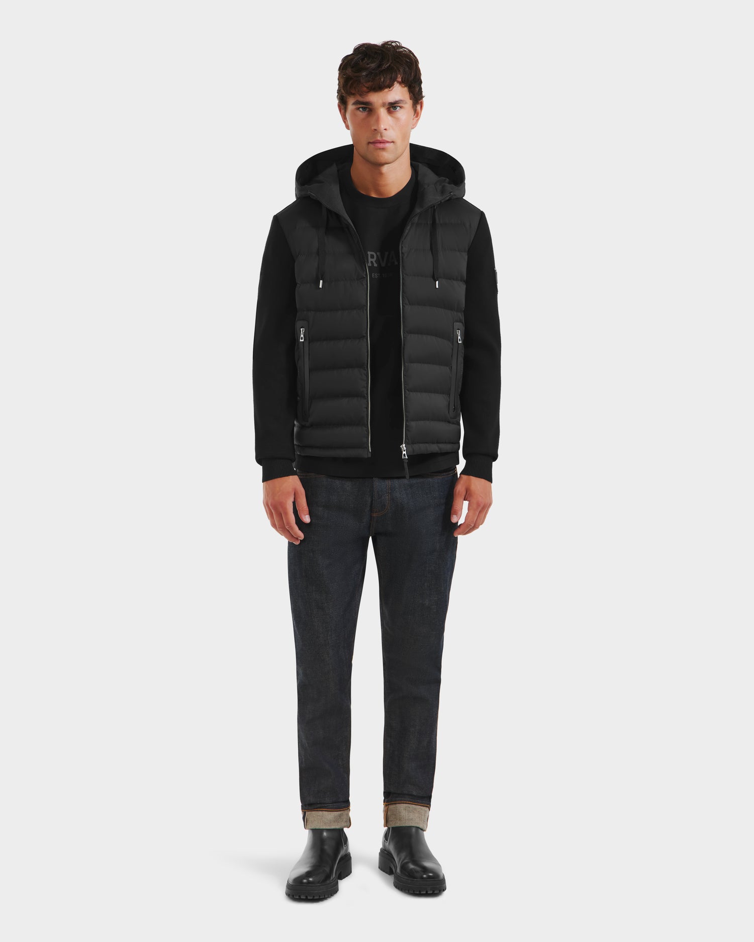 A man stands facing forward against a white background, wearing the Rudsak RIVER HV - H125503 Black limited edition Harvard x RUDSAK hybrid hooded down jacket, dark rolled-cuff jeans, and black shoes.