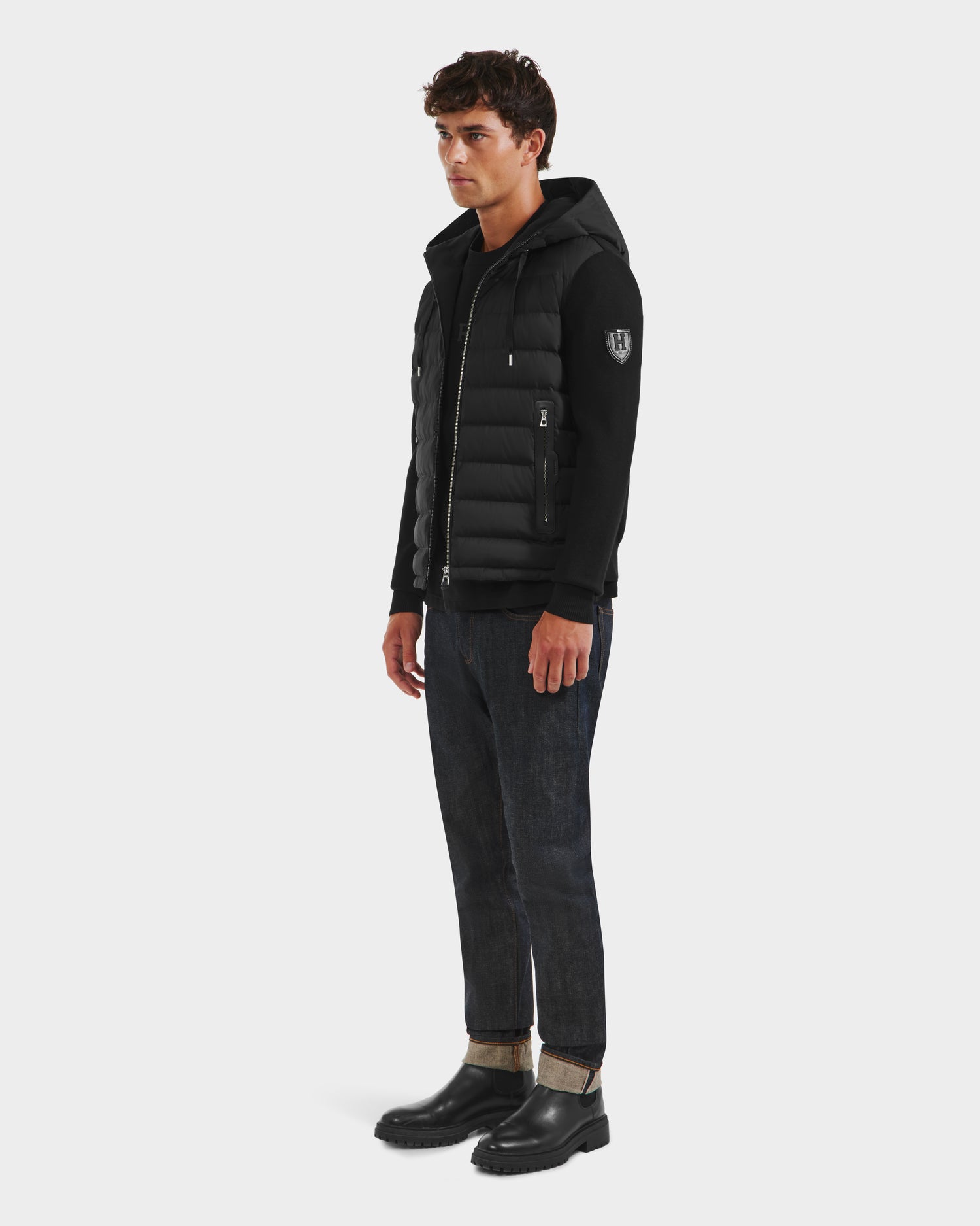 A man faces slightly sideways wearing the Rudsak RIVER HV - H125503 Black hybrid hooded down jacket, paired with dark cuffed jeans and black shoes, set against a plain white background.