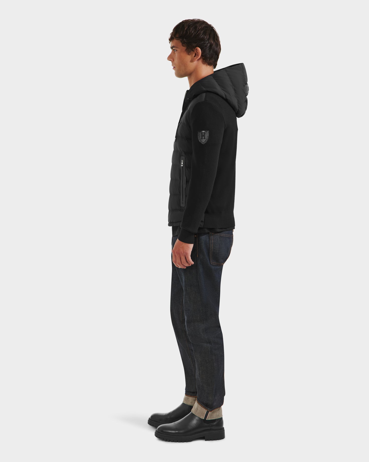 A man stands in profile wearing the limited edition Rudsak RIVER HV - H125503 Black hybrid hooded down jacket, dark cuffed jeans, and black boots against a plain white background.