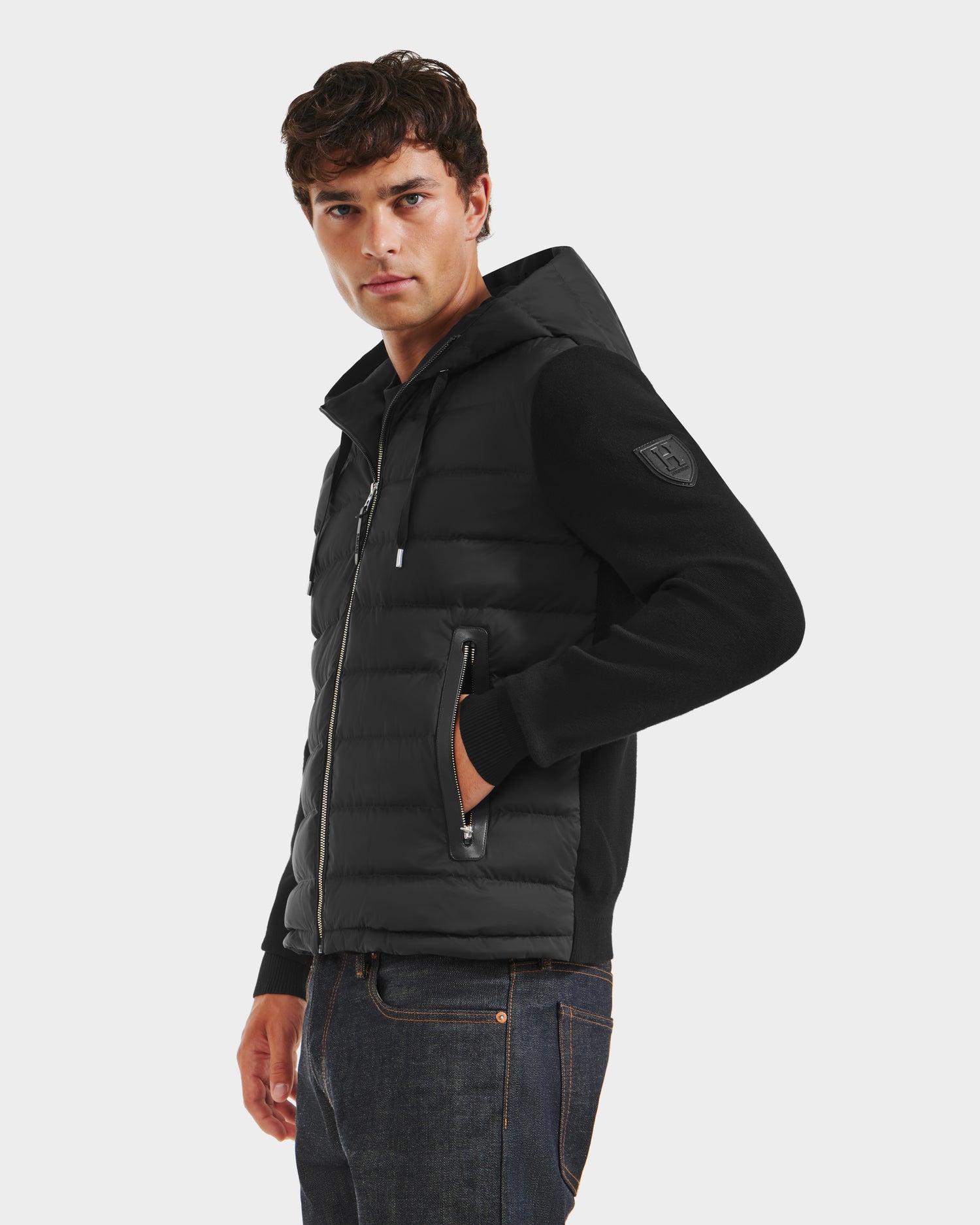 A young man stands against a plain white background, wearing the Rudsak RIVER HV - H125503 Black hybrid hooded down jacket with zippered pockets and dark jeans, facing slightly to the side with one hand in his pocket.