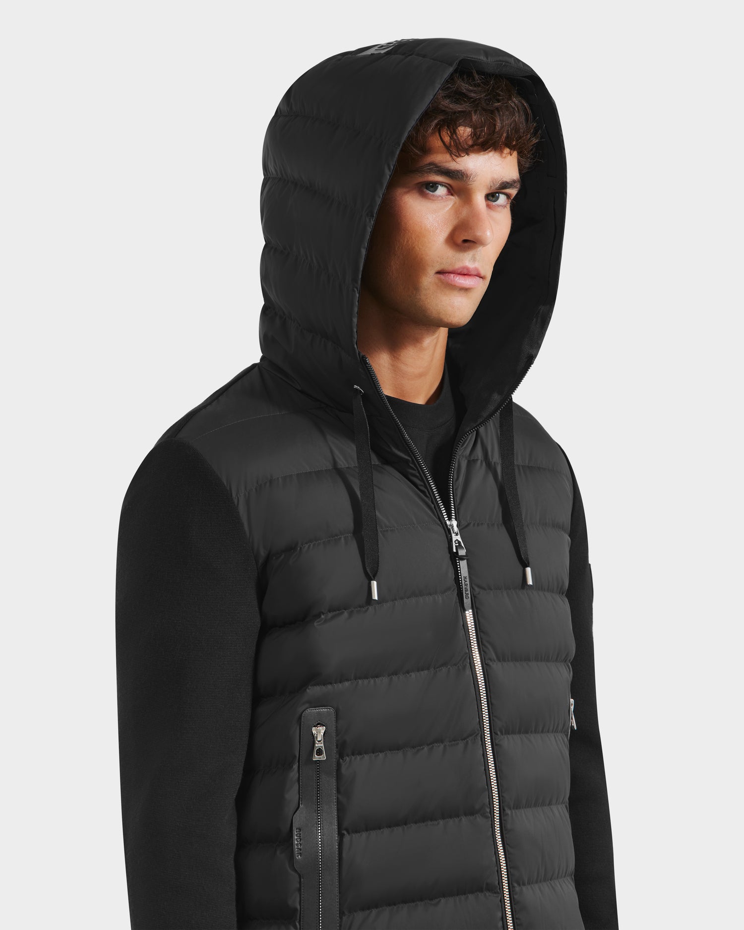 A young man with short brown hair wears the Rudsak RIVER HV - H125503 Black hybrid hooded down jacket over a black shirt, gazing seriously at the camera against a plain light background.
