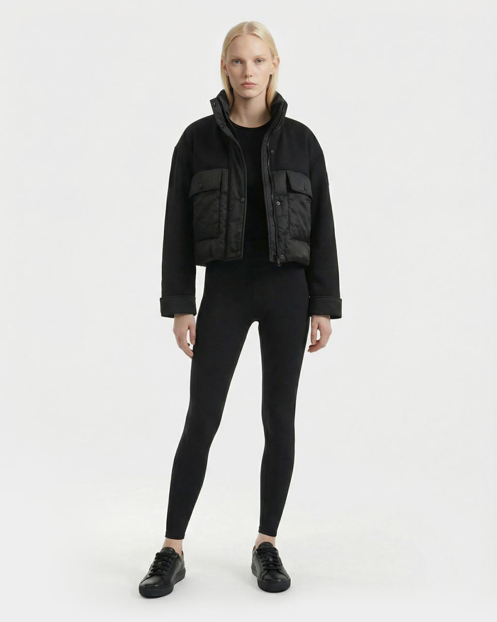 A person with long blonde hair faces forward, wearing the Rudsak ROME - 8126028 Black mixed-media cropped jacket with large front pockets, paired with black leggings and sneakers, against a plain white background.