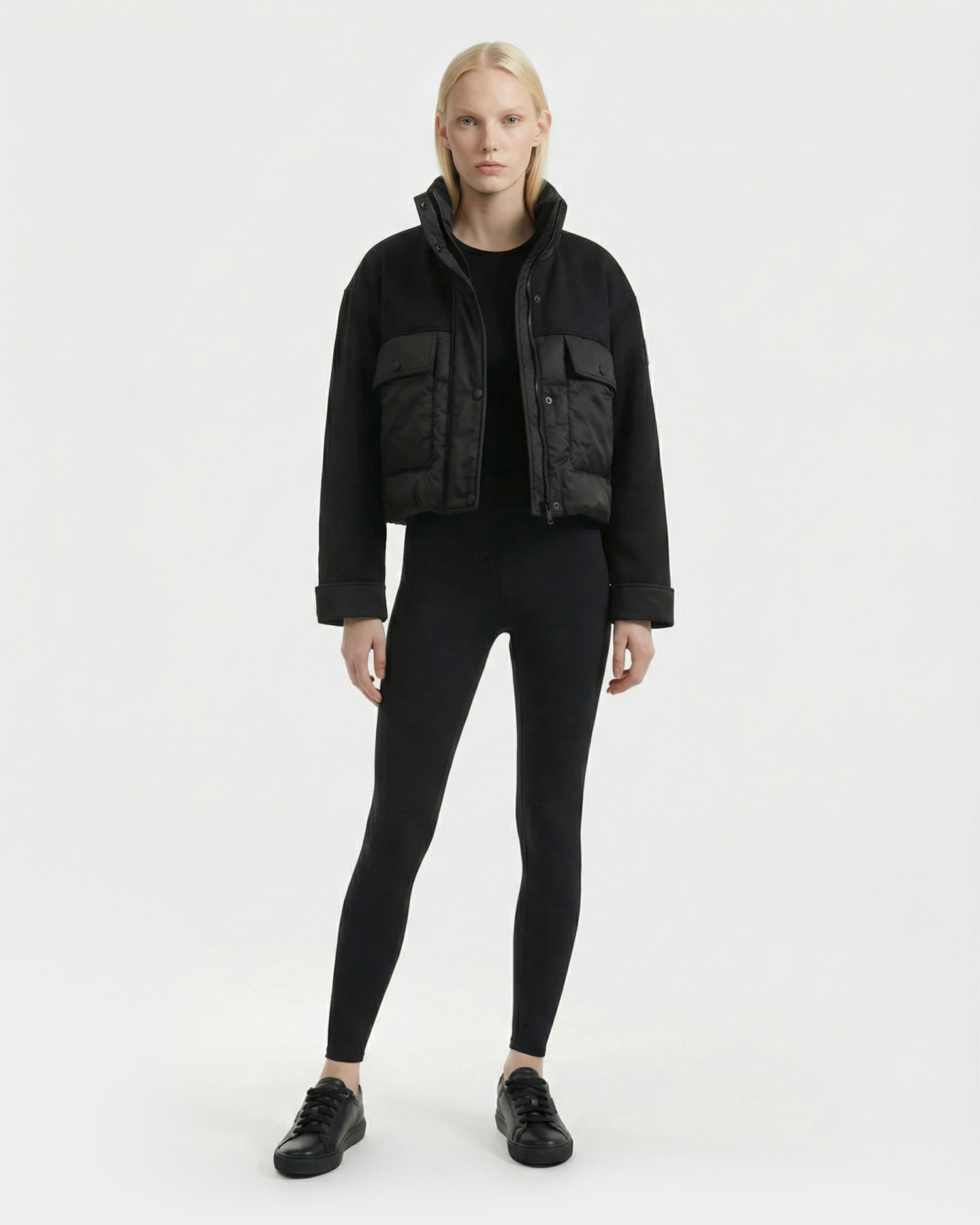 A person with long blonde hair faces forward, wearing the Rudsak ROME - 8126028 Black mixed-media cropped jacket with large front pockets, paired with black leggings and sneakers, against a plain white background.