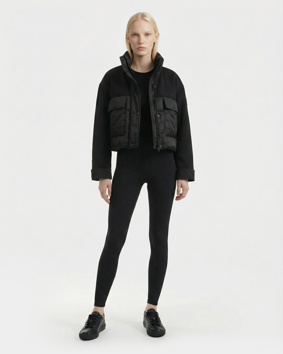 A person with long blonde hair faces forward, wearing the Rudsak ROME - 8126028 Black mixed-media cropped jacket with large front pockets, paired with black leggings and sneakers, against a plain white background.