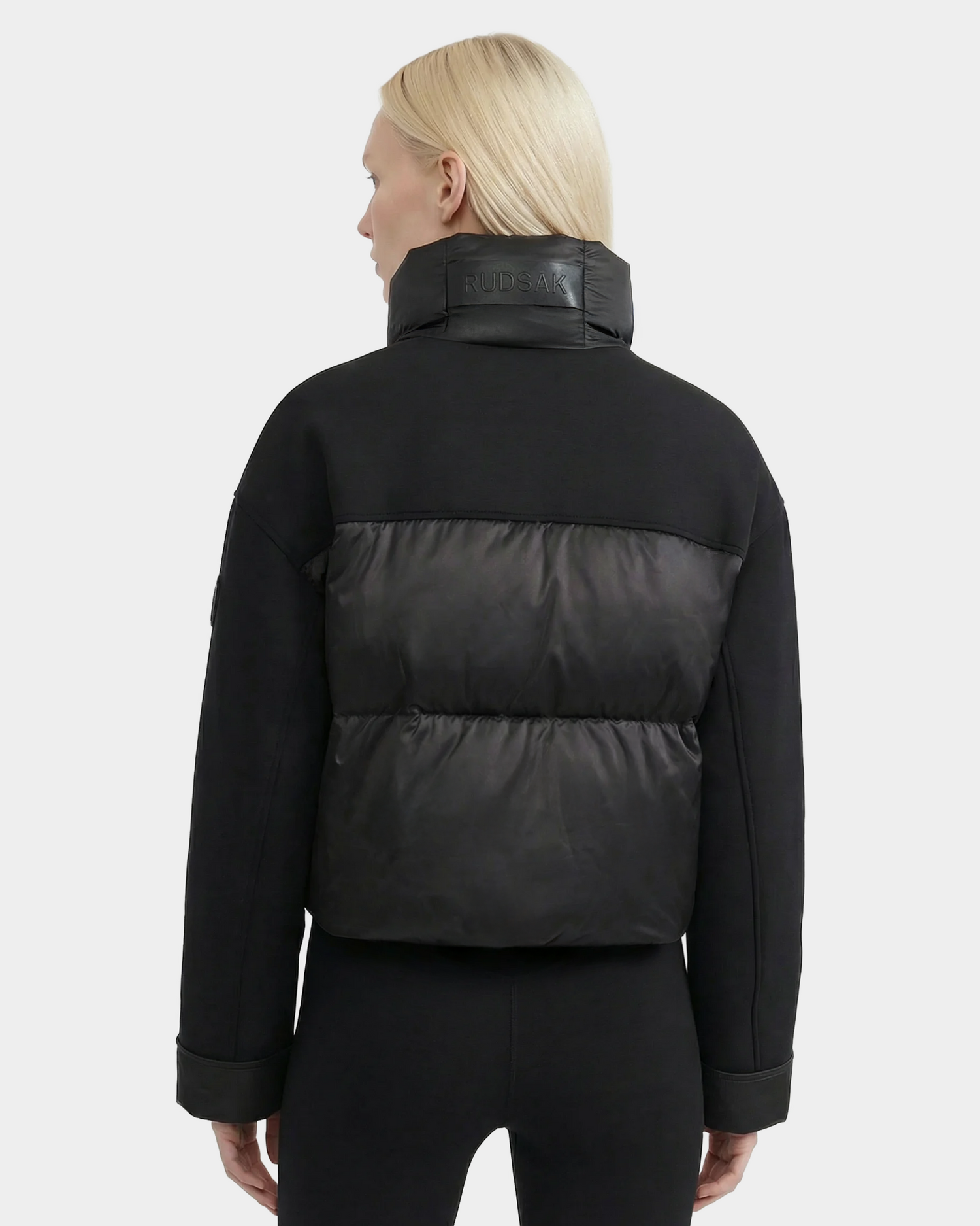 A person with straight light blonde hair, dressed in the Rudsak ROME - 8126028 Black quilted down-filled jacket and black pants, is shown from behind against a plain light gray background.