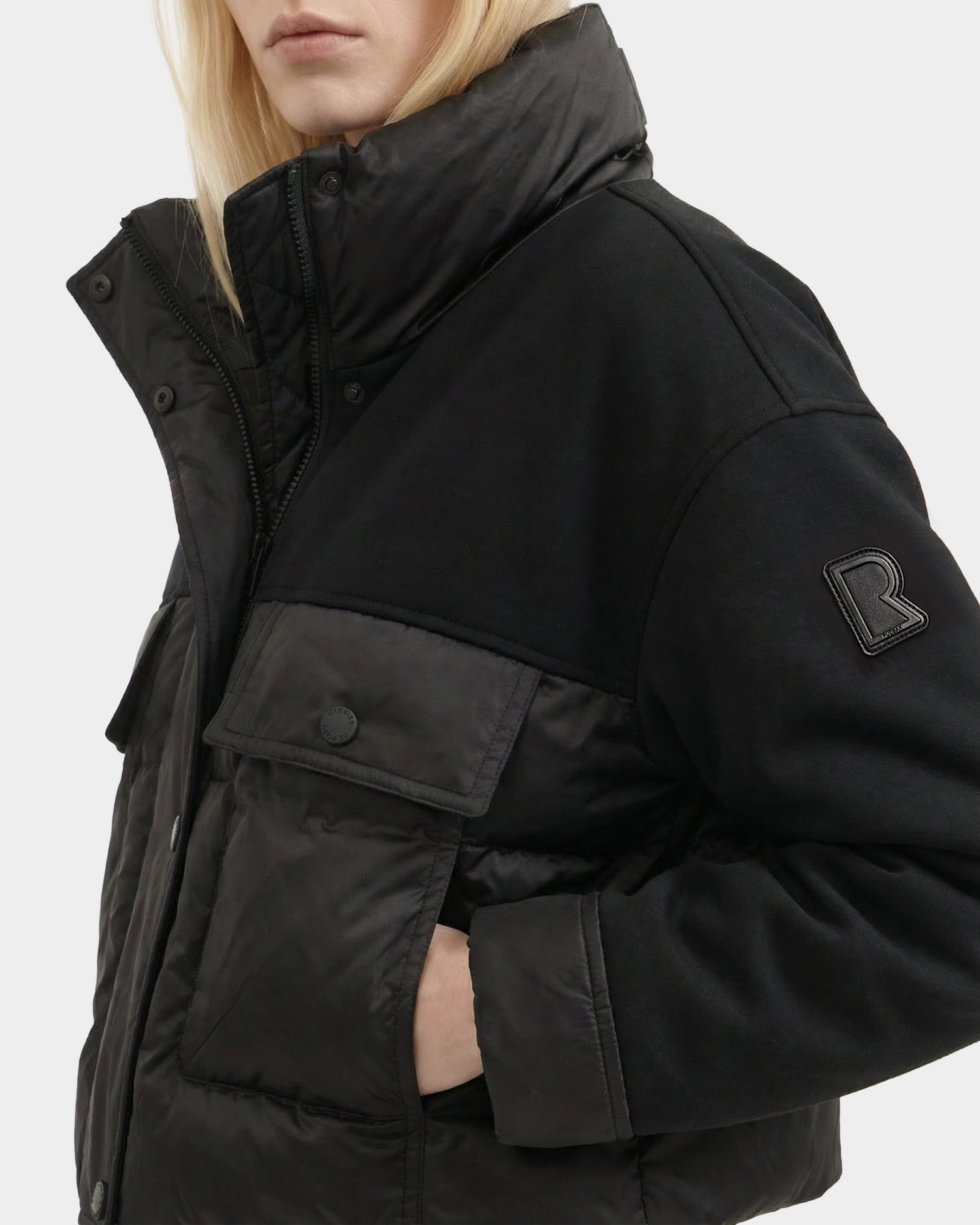 A person with long blonde hair wears the ROME - 8126028 Black quilted down puffer jacket by Rudsak, featuring a high collar, large buttons, front pockets, an R patch on the sleeve, and their hand in a pocket.