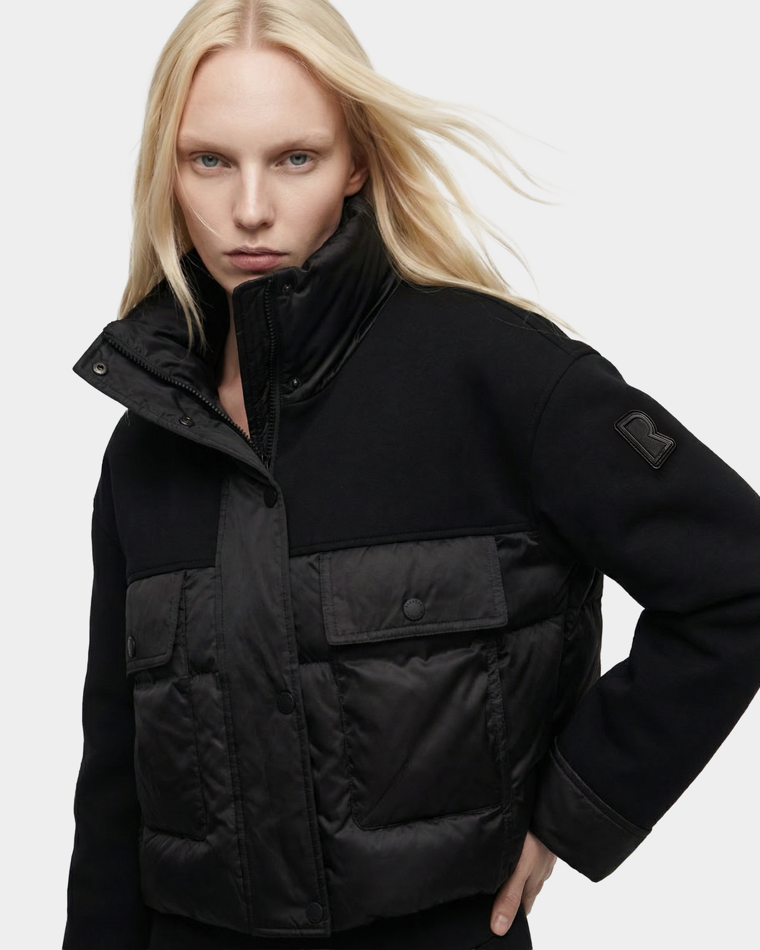 A woman with long blonde hair models the Rudsak ROME - 8126028 Black quilted down jacket, featuring large front pockets and a high collar, posing with one hand on her hip against a plain light background.