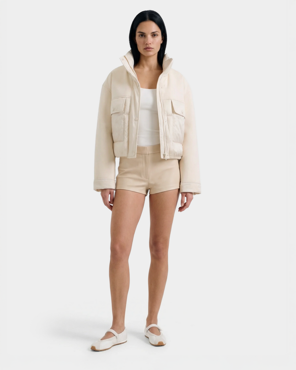 A woman with straight dark hair and a neutral expression stands against a plain background, wearing the Rudsak ROME - 8126028 Iceland beige mixed-media jacket for lightweight warmth, paired with a white tank top, beige shorts, and white flats.