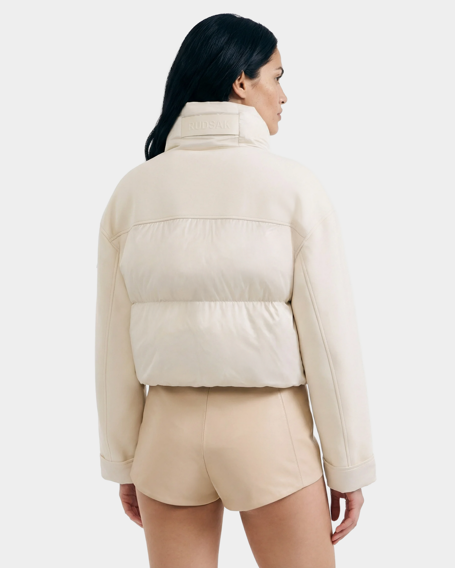 A woman with long dark hair, seen from behind, wears the Rudsak ROME - 8126028 Iceland light beige quilted down-filled puffer jacket and matching high-waisted shorts, offering lightweight warmth against a plain light background.