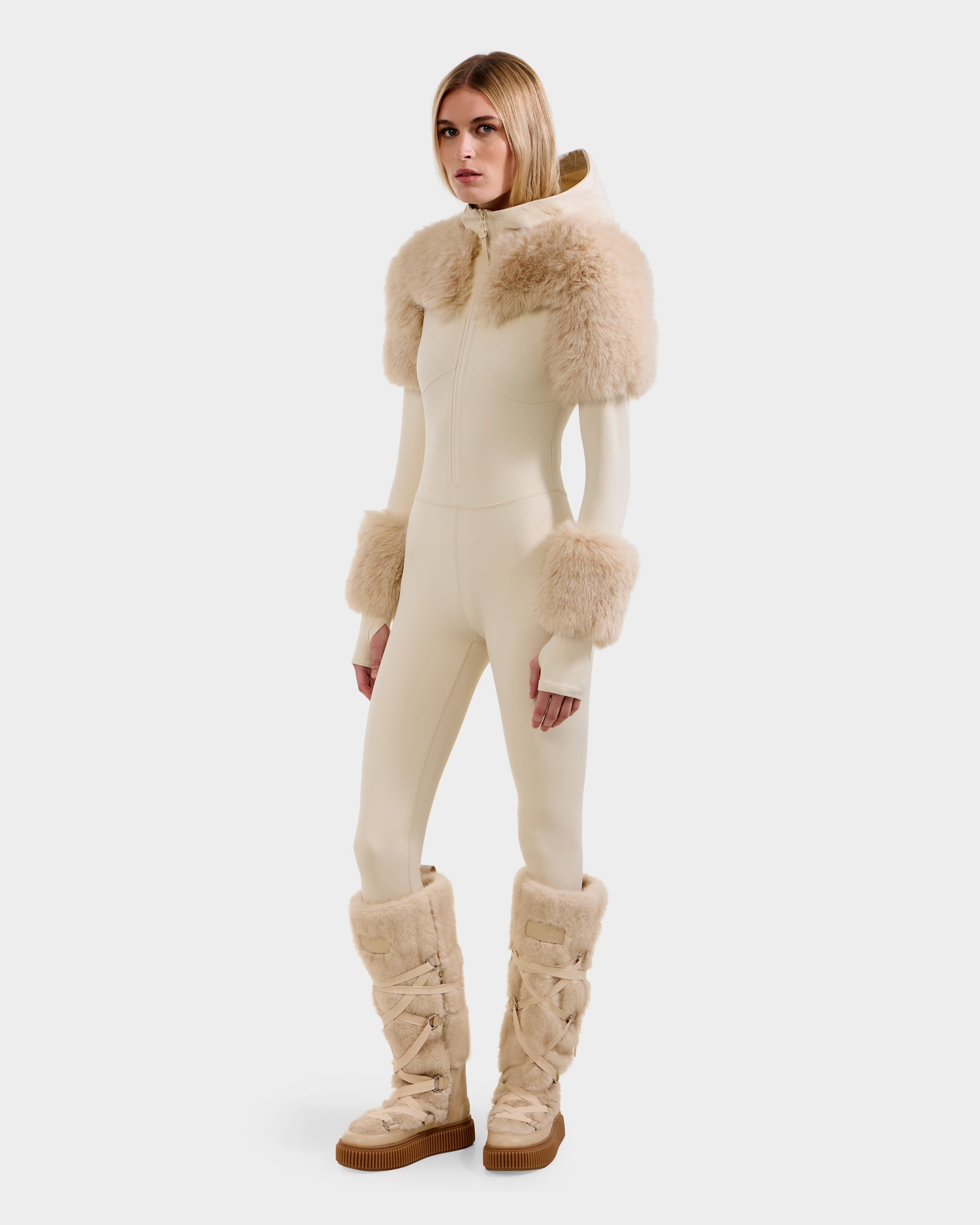 A woman models the Rudsak MYLA - 8525671 Vanilla, a cream jumpsuit with matching faux fur on the shoulders, cuffs, and tall boots. Featuring a high collar and front zipper, she poses chicly against a plain white background.