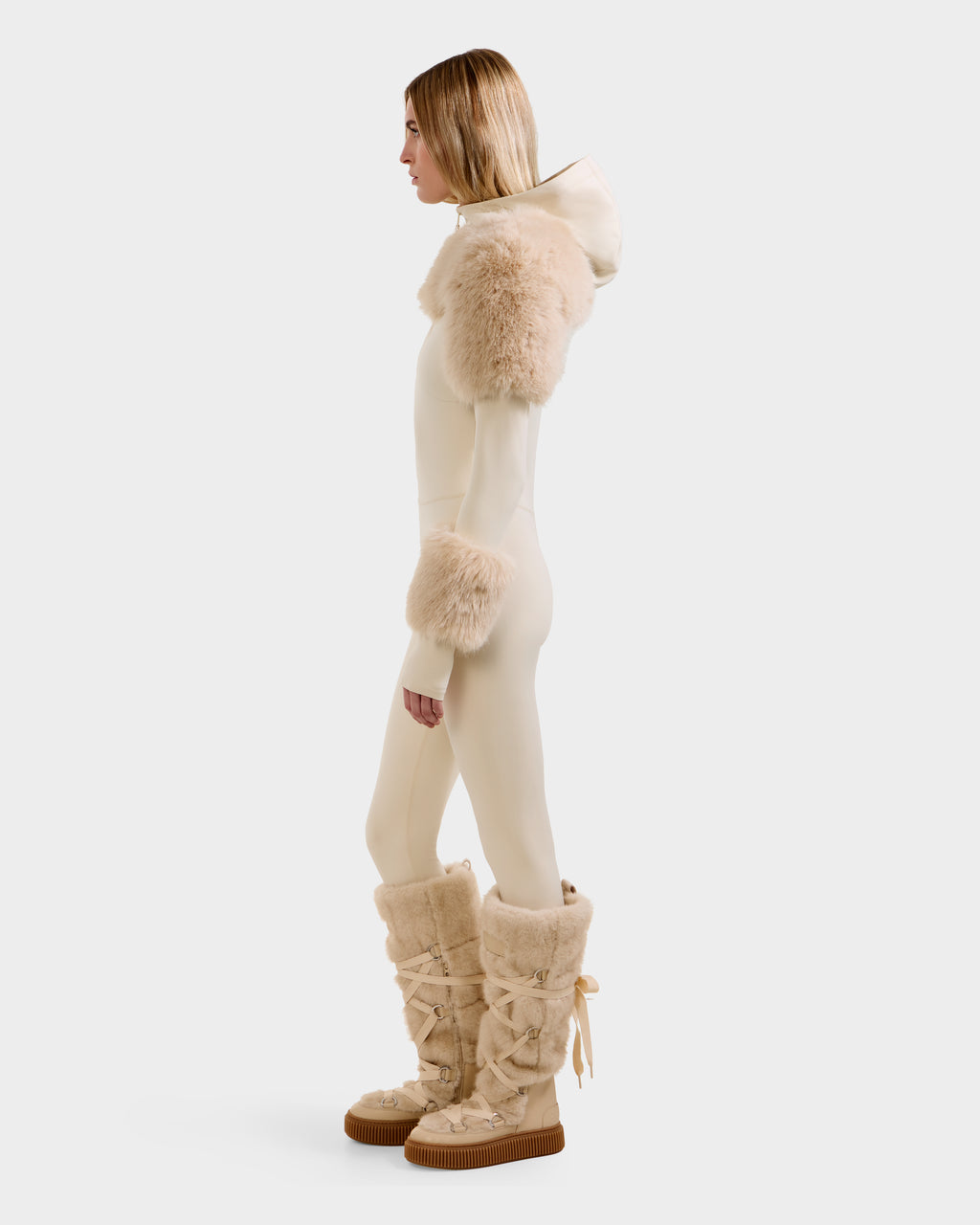 A person stands in profile wearing the Rudsak MYLA - 8525671 Vanilla women’s bodysuit with a hood and plush faux fur trim, plus matching tall fur boots—a chic après-ski winter look against a plain, light background.