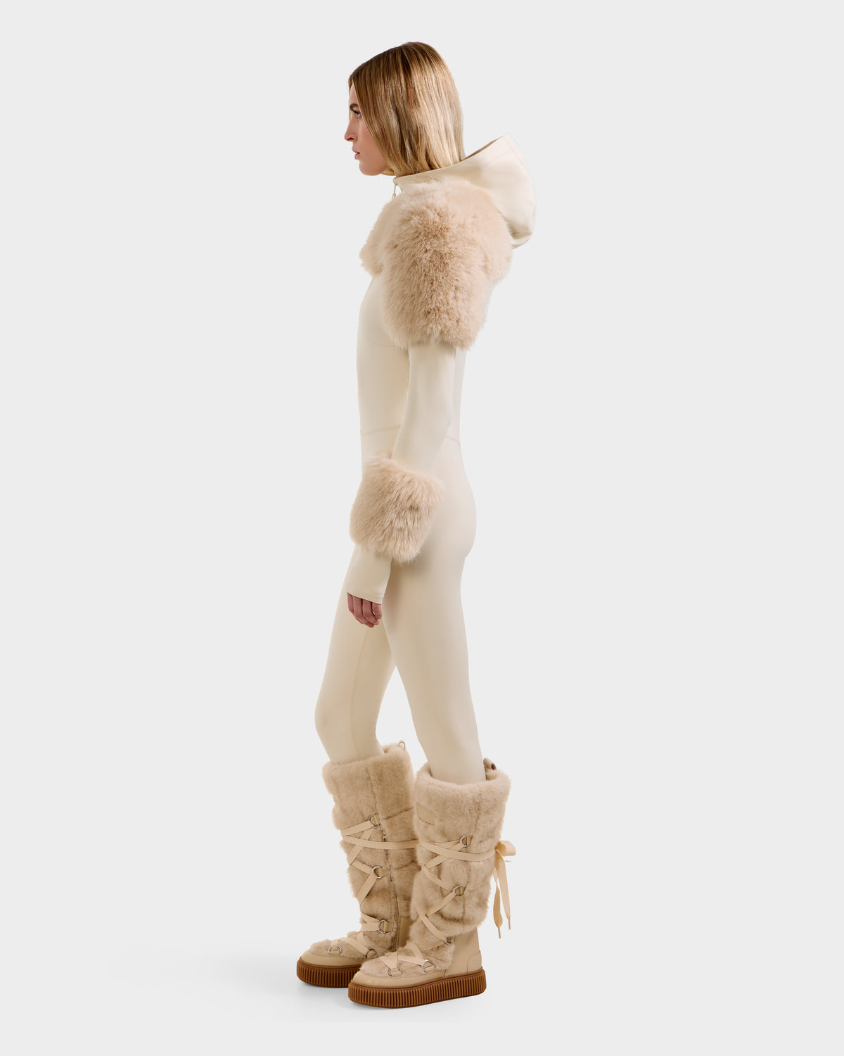 A person stands in profile wearing the Rudsak MYLA - 8525671 Vanilla women’s bodysuit with a hood and plush faux fur trim, plus matching tall fur boots—a chic après-ski winter look against a plain, light background.