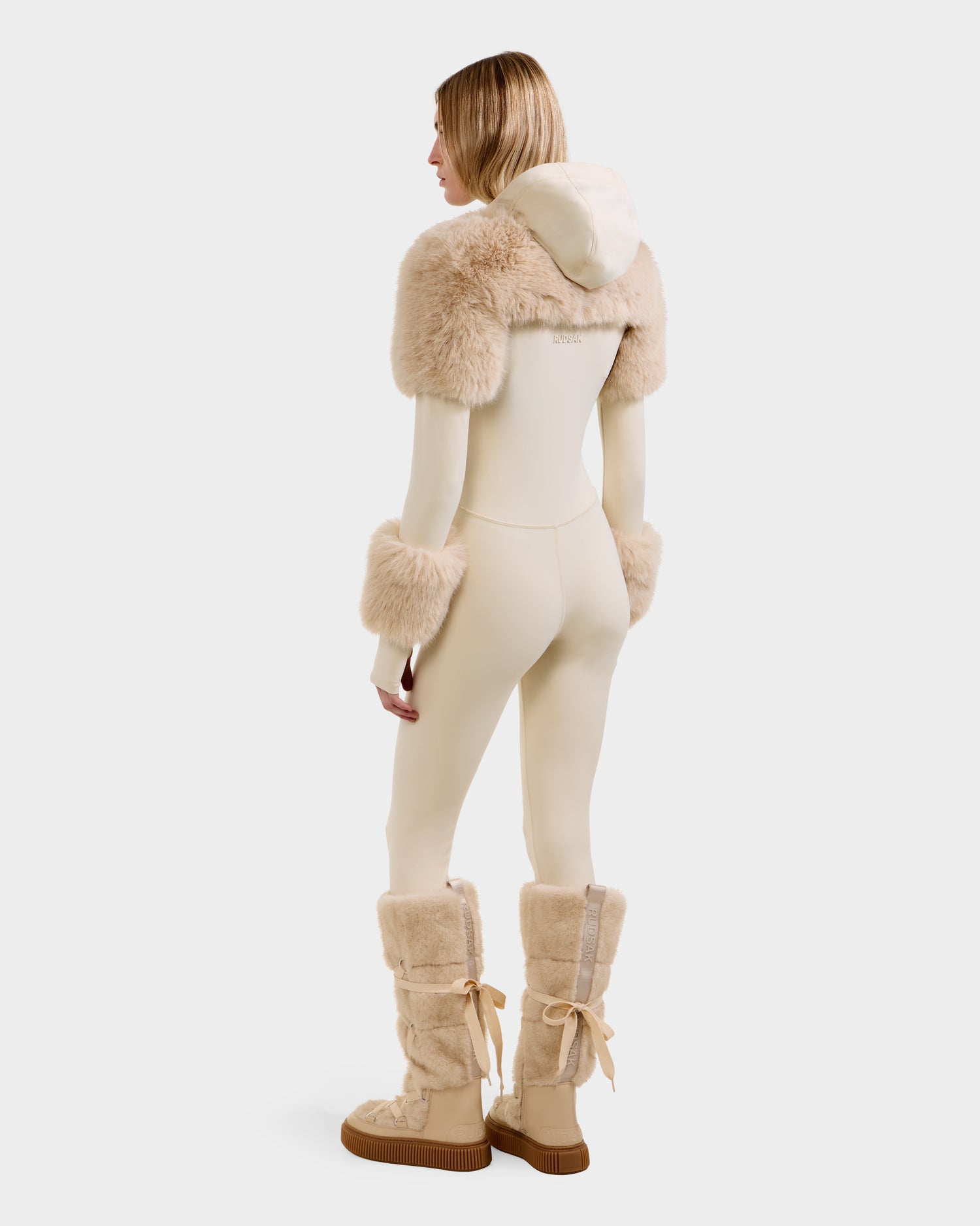A woman faces away, wearing the Rudsak MYLA - 8525671 Vanilla fitted bodysuit with furry beige accents on the cuffs, shoulders, and hood, plus matching furry boots—an après-ski winter look—set against a plain white background.