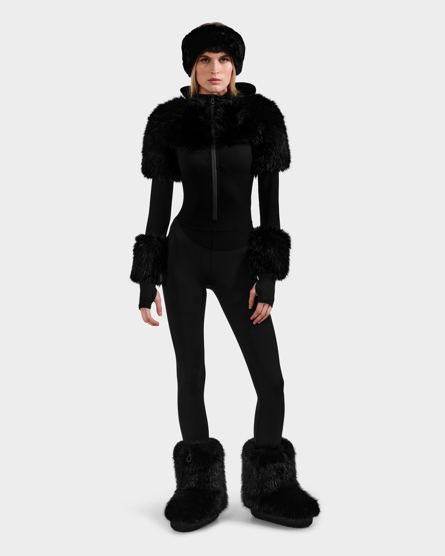 A person faces forward in the Rudsak MYLA - 8525671 Black fitted women’s bodysuit, featuring faux fur trim on shoulders, wrists, boots, and a matching fur hat—ideal for après-ski—against a plain white background.