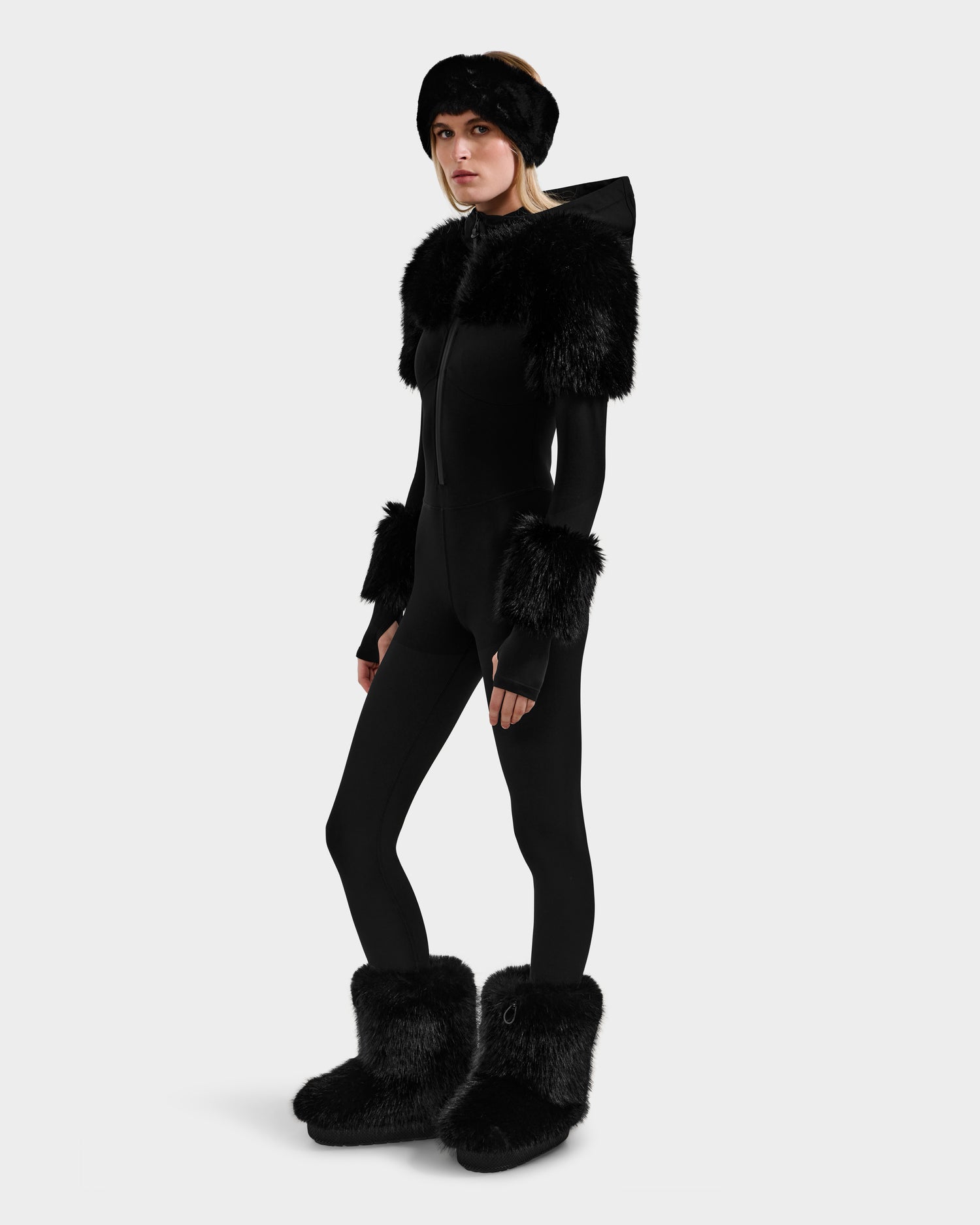 A person wears the Rudsak MYLA - 8525671 Black women’s bodysuit, featuring black faux fur on the shoulders, wrists, boots, and headband, standing slightly turned against a plain light background.