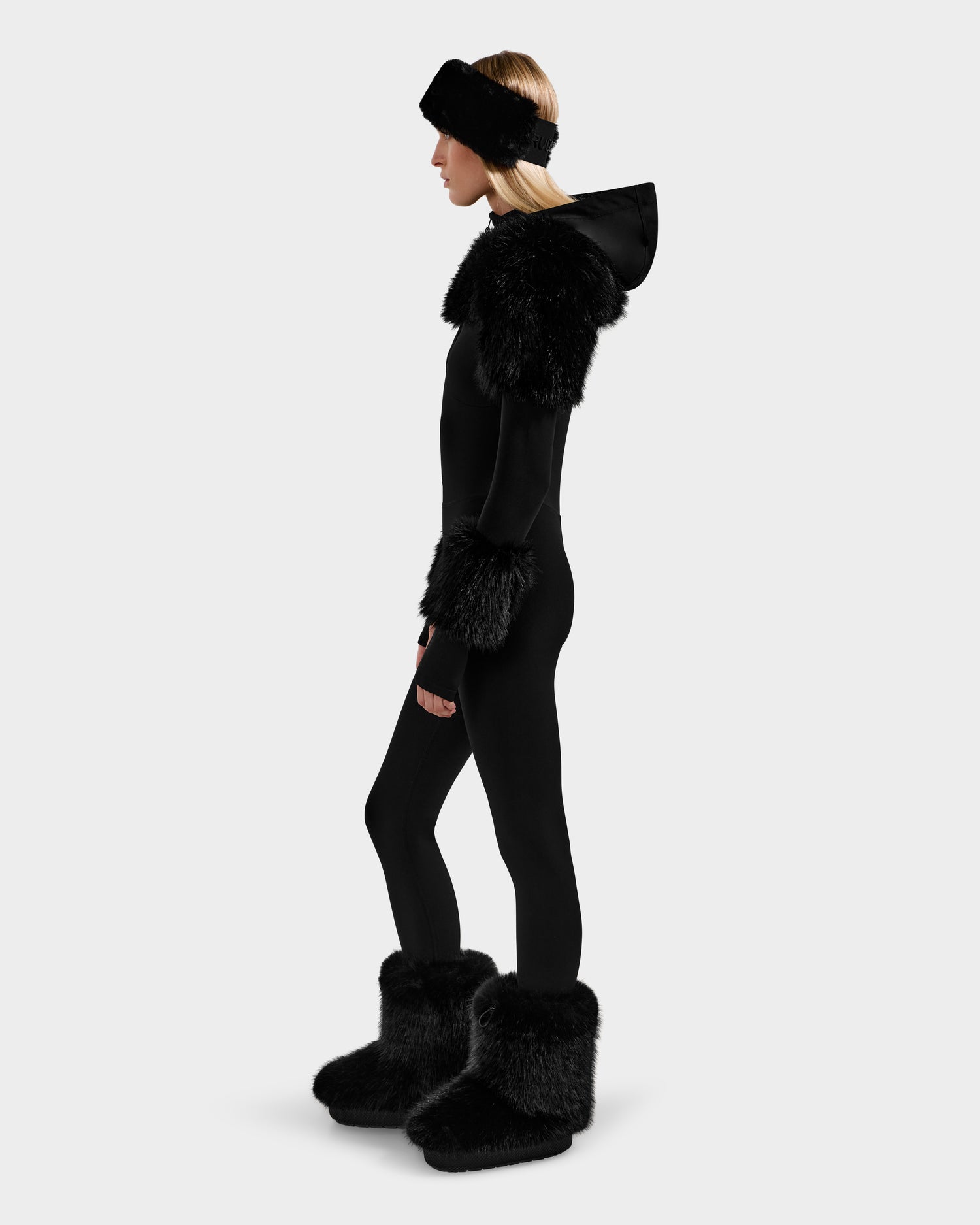 A woman in profile wears the Rudsak MYLA - 8525671 Black fitted bodysuit with faux fur cuffs, shoulder accents, boots, and a matching headband against a plain light background.