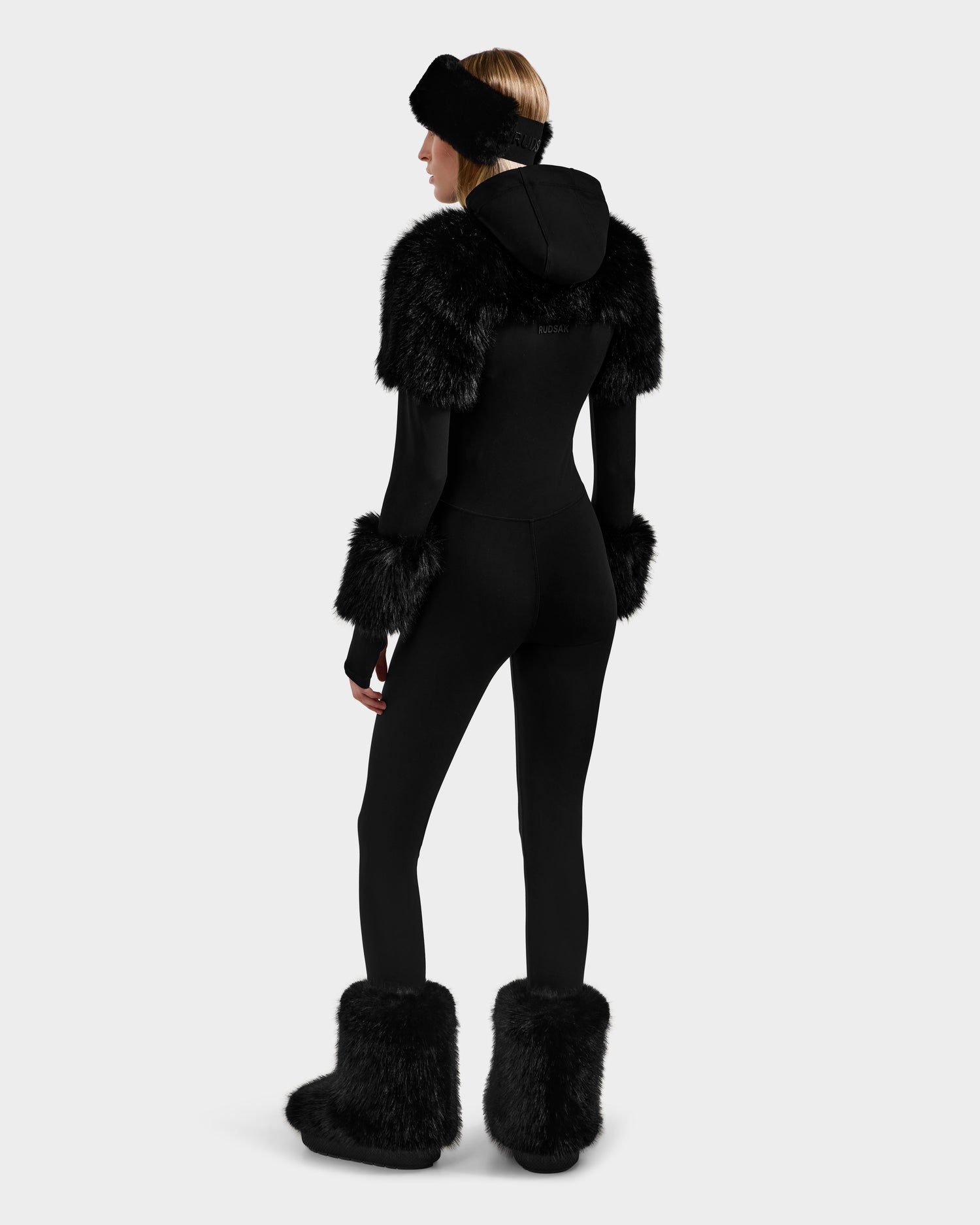 A person faces away, wearing Rudsak's MYLA - 8525671 Black fitted women’s bodysuit with black faux fur on shoulders, cuffs, boots, and a matching headband—ideal for après-ski. Background is plain light gray.