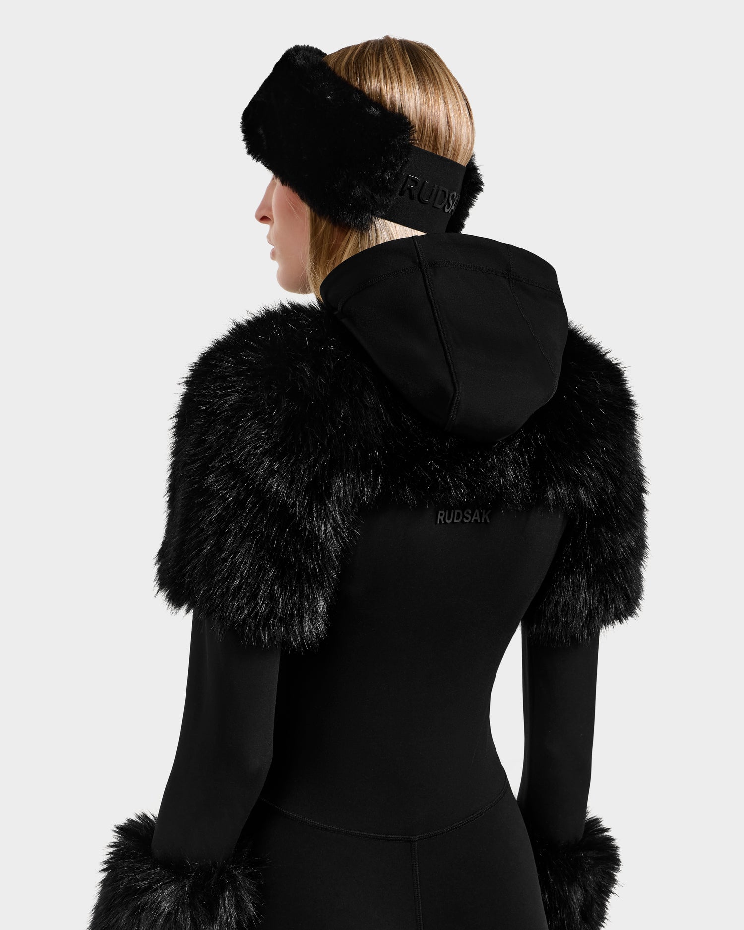 Styled for après-ski, a person is seen from the back in the RUDSAK MYLA - 8525671 Black jacket with faux fur-trimmed hood, matching fur cuffs, and a black Rudsak headband, set against a light background.