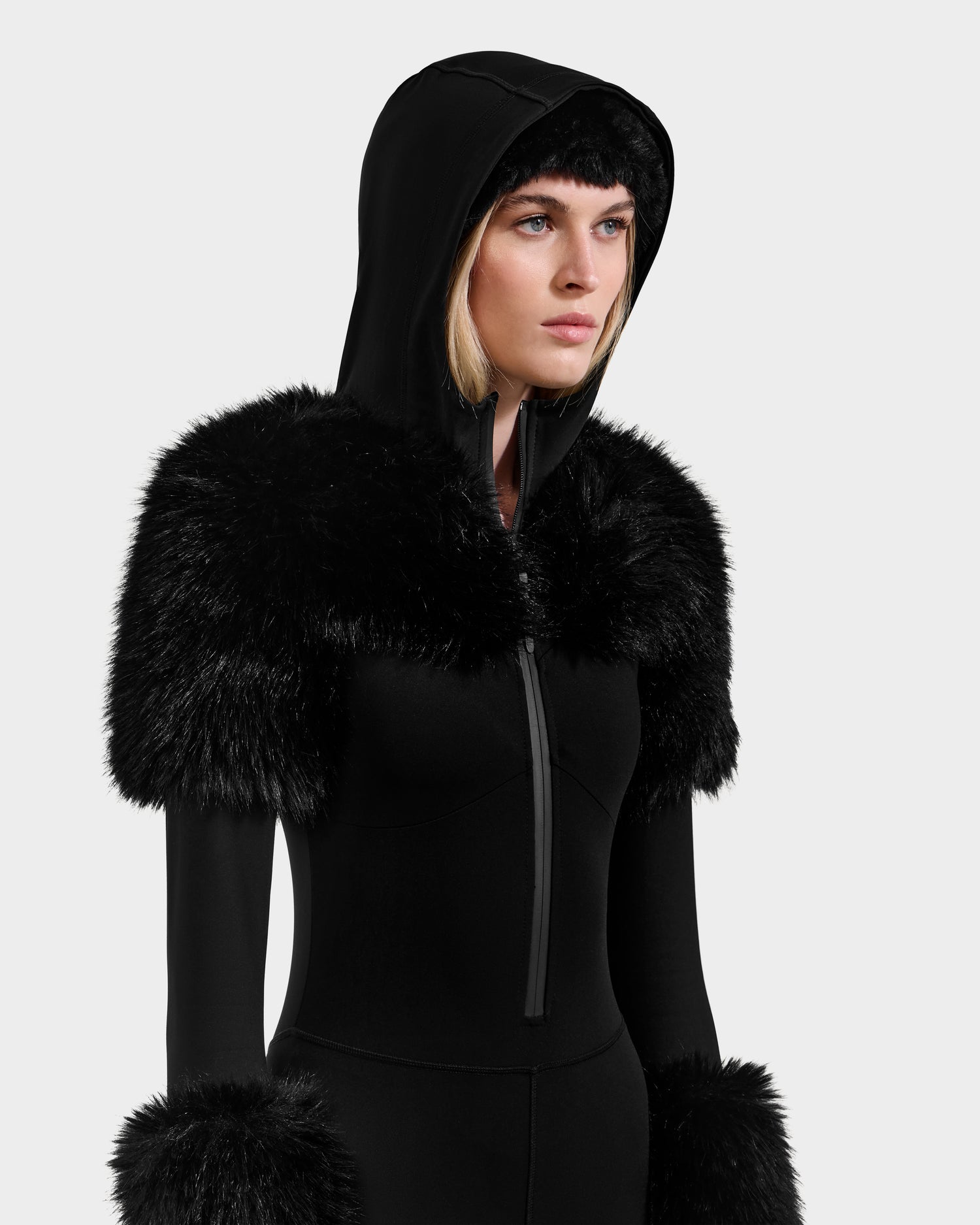 A woman wears the Rudsak MYLA - 8525671 Black hooded après-ski outfit with faux fur on the shoulders and cuffs, facing right against a plain light gray background.