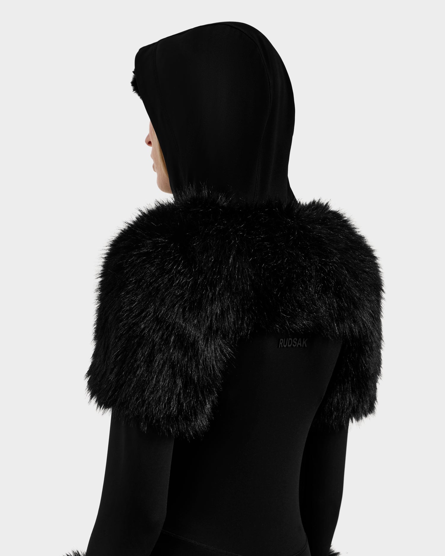 A person wears the Rudsak MYLA - 8525671 Black coat, featuring a large, fluffy faux fur collar and hood, viewed from the back and side, adding après-ski style against a plain white background.