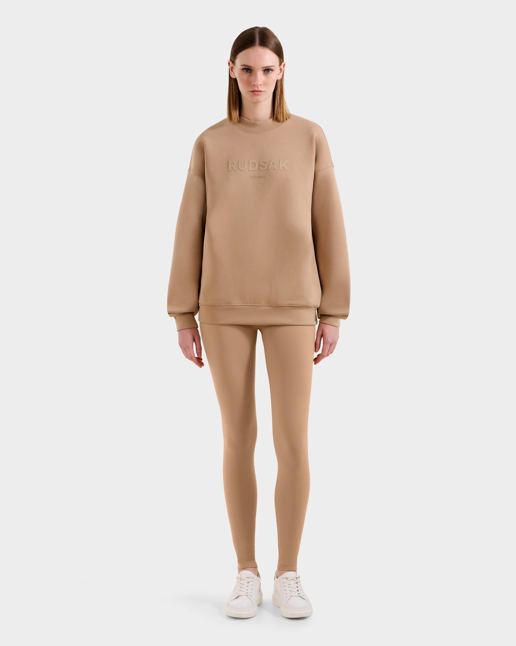 A woman faces forward in the Rudsak BRYN - 9524007 Almond W cotton crewneck sweatshirt with embossed branding, paired with matching leggings and white sneakers—an effortlessly timeless look against a plain white background.