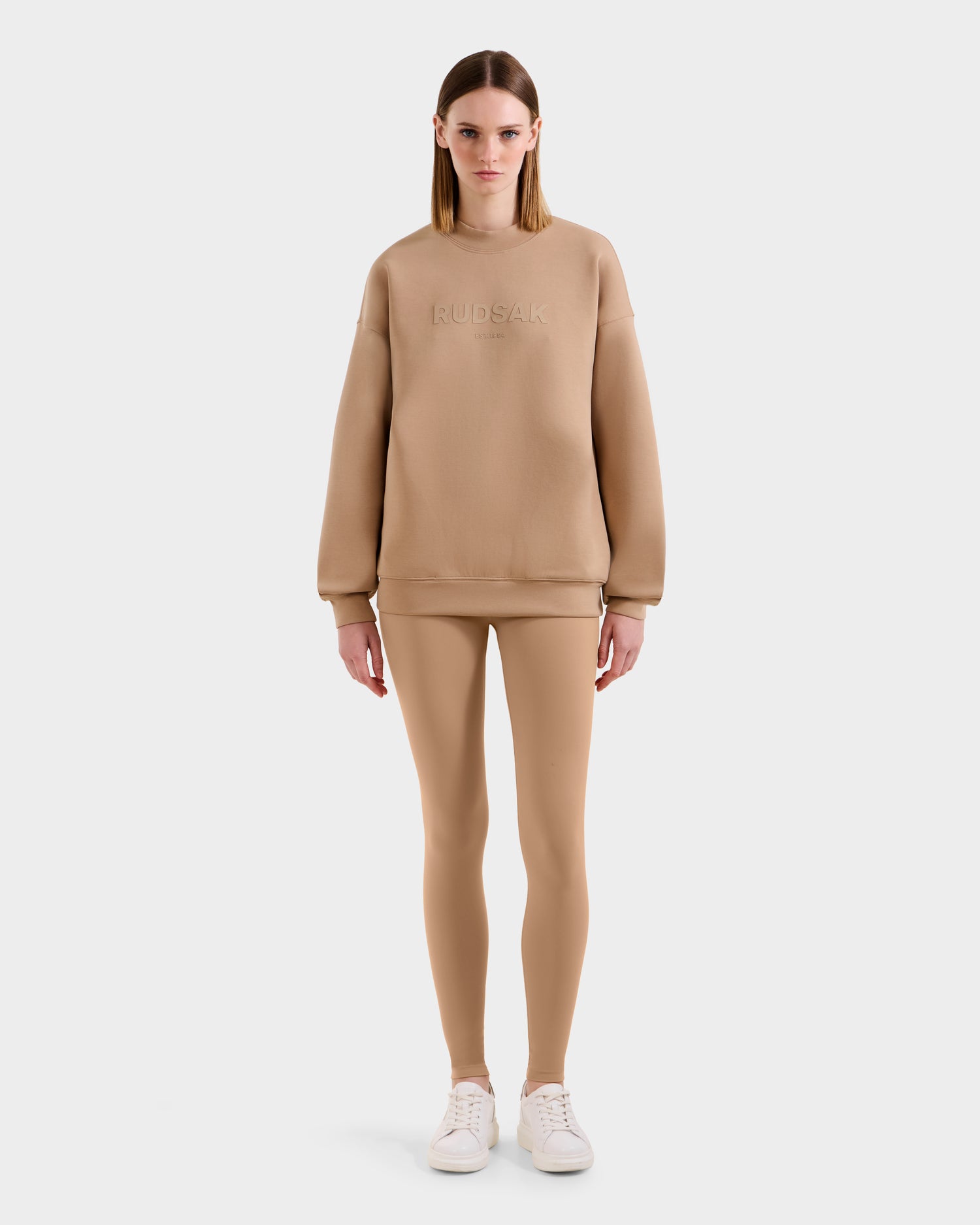 A woman faces forward in the Rudsak BRYN - 9524007 Almond W cotton crewneck sweatshirt with embossed branding, paired with matching leggings and white sneakers—an effortlessly timeless look against a plain white background.