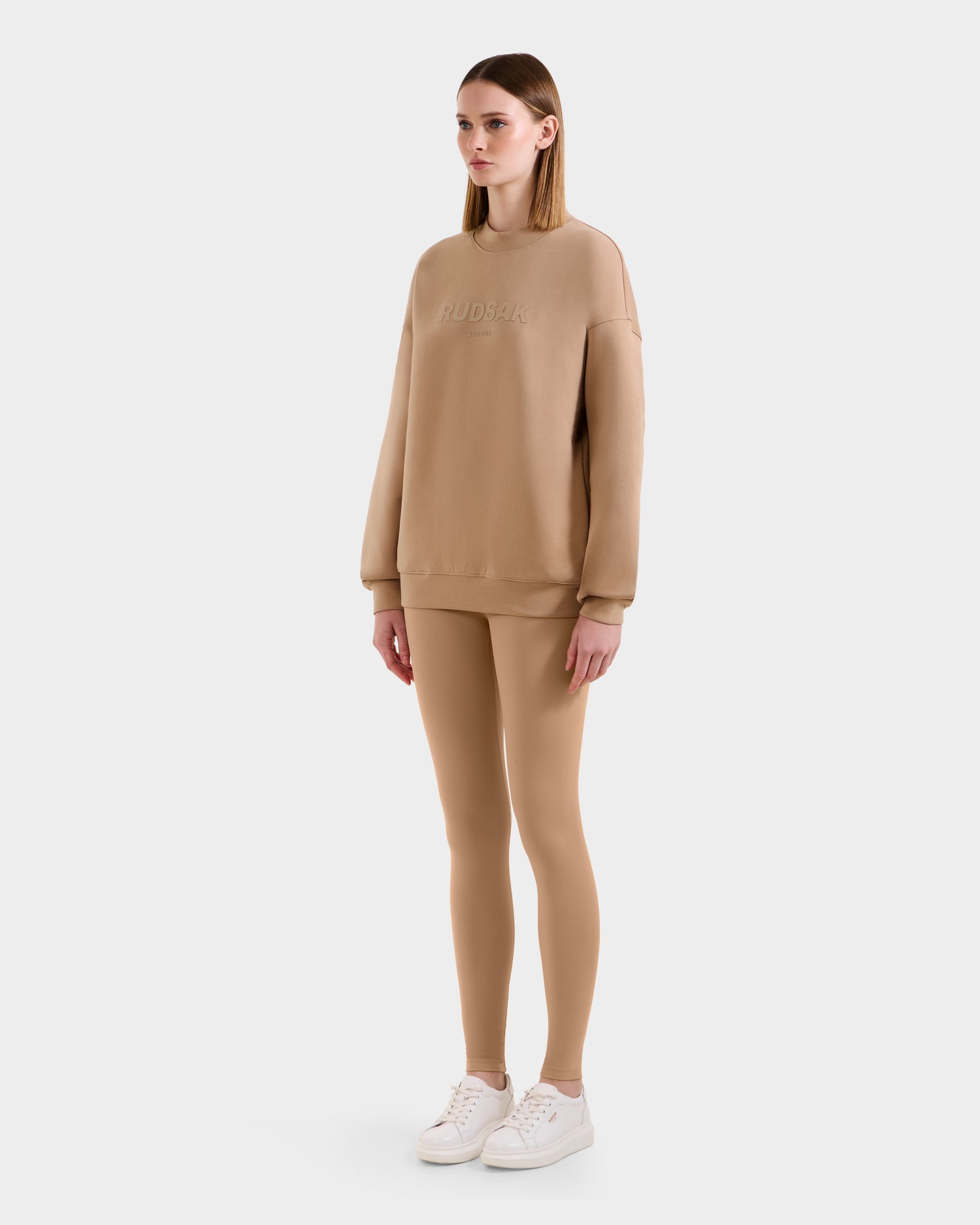 A woman wears the Rudsak BRYN - 9524007 Almond W cotton crewneck sweatshirt and matching leggings set with white sneakers. The oversized fit and subtle text create a timeless look as she faces slightly right, expression neutral.