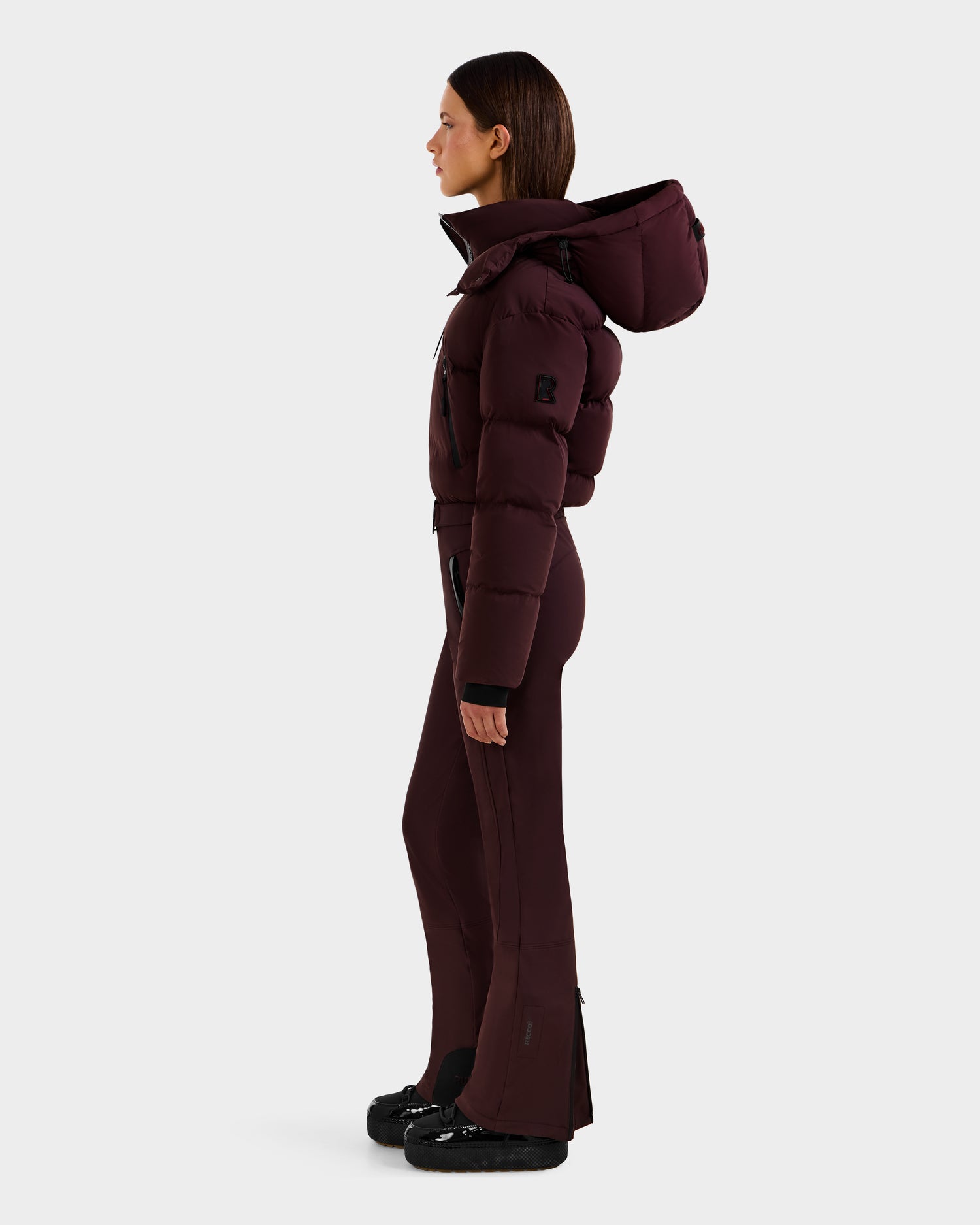 A woman stands in profile against a light gray background wearing the Rudsak DINARA - 8125600 Bordeaux ski suit with a large hood and fitted pants, paired with black shoes.