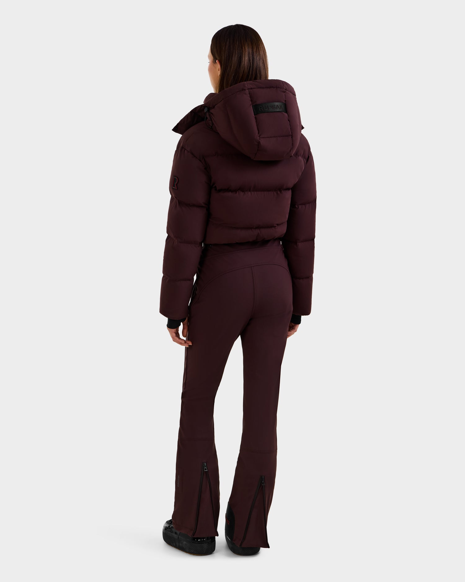 A person faces away, wearing the Rudsak DINARA - 8125600 Bordeaux women’s ski suit with hood and zippered flares, plus black shoes, set against a plain light gray background.
