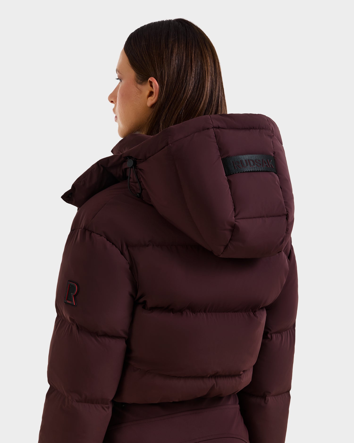 A woman with straight brown hair is seen from the back wearing the Rudsak DINARA - 8125600 Bordeaux puffer jacket, featuring a black Rudsak label on the hood and an R patch on the sleeve—ideal for layering over your women’s ski suit.